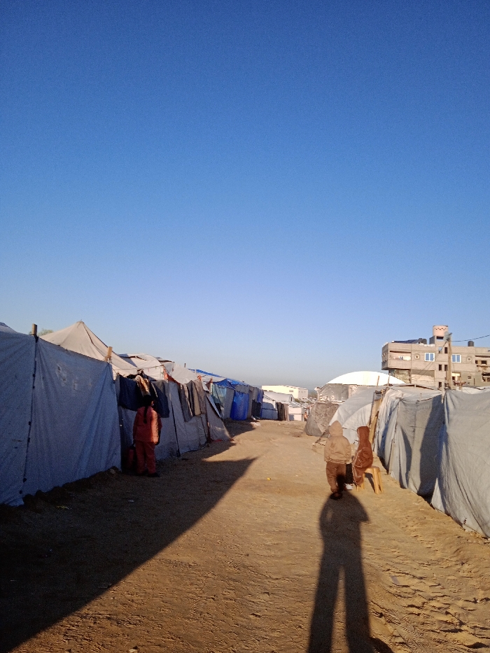 The image presents a ground-level view of a narrow, unpaved dirt path traversing an informal settlement in Khan Yunis, Palestinian Territory, under a clear, bright blue sky. On both the left and right sides of the path, rows of closely spaced makeshift shelters are constructed from white and blue tarpaulins, fabrics, and various support poles. On the left side, clothing items are observed hanging on the exterior of several shelters. An individual in a red or orange garment stands facing one of these shelters on the left. Further down the path on the right, two figures are walking away from the viewer; one is wearing a light brown hooded top, and the other, adjacent to them, is next to a small wooden stool. Long shadows, including a distinct human shadow in the foreground, stretch across the path, indicating a low sun angle. In the distant background, a multi-story concrete building with windows and a rooftop water tank is visible on the right, alongside a large white dome-like structure and additional informal dwellings.