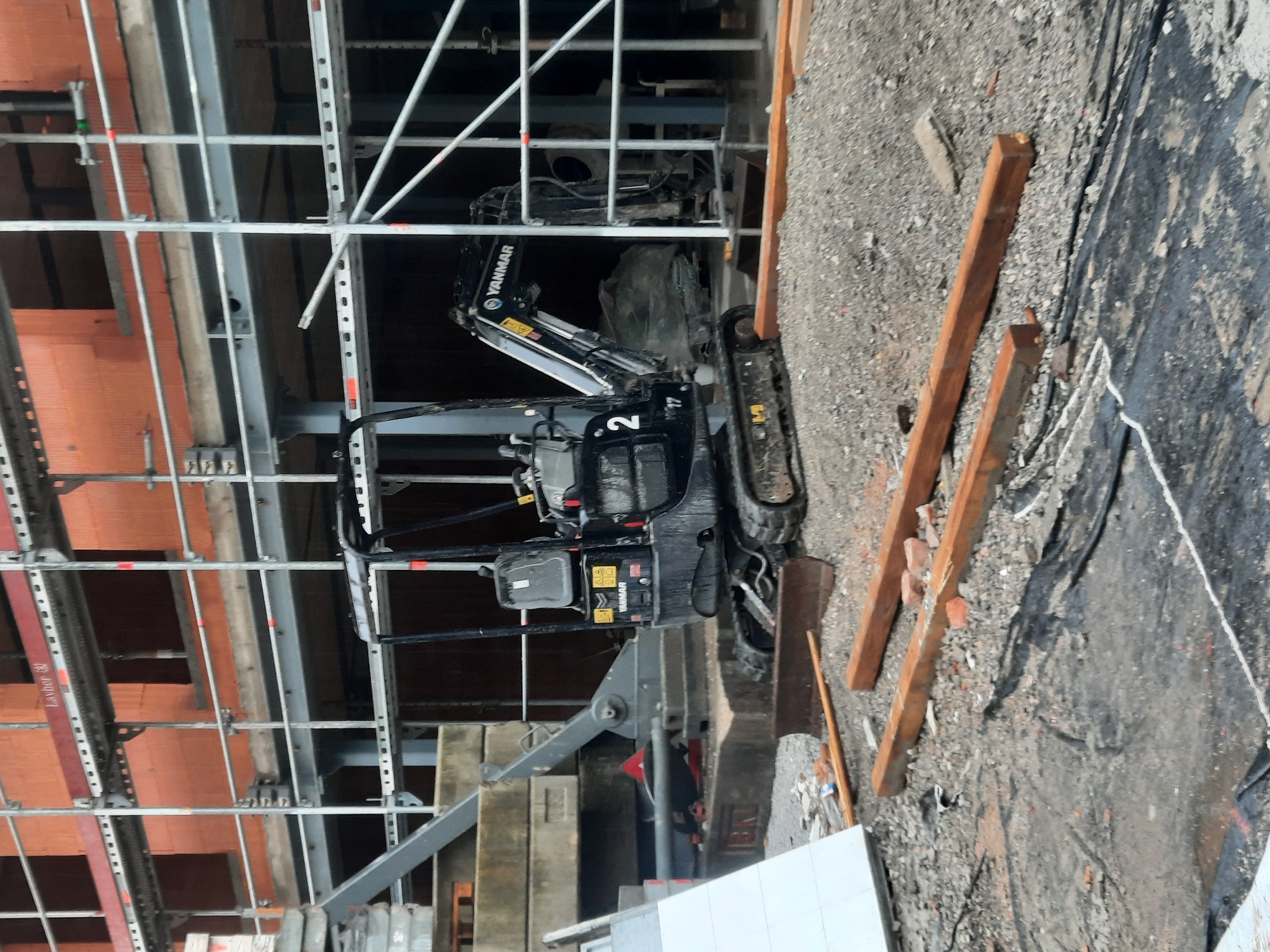 This is a medium-distance, eye-level shot of a construction site in Rosenheim, Germany. The foreground is cluttered with debris including gravel, wooden planks, and what appears to be a tarpaulin. A small, black Yanmar excavator is the central focus, positioned on the gravel. It has the number "2" visible on its side. Behind the excavator, scaffolding with orange brick walls is visible. To the left of the excavator, there are large concrete blocks. The overall scene depicts an active construction zone.