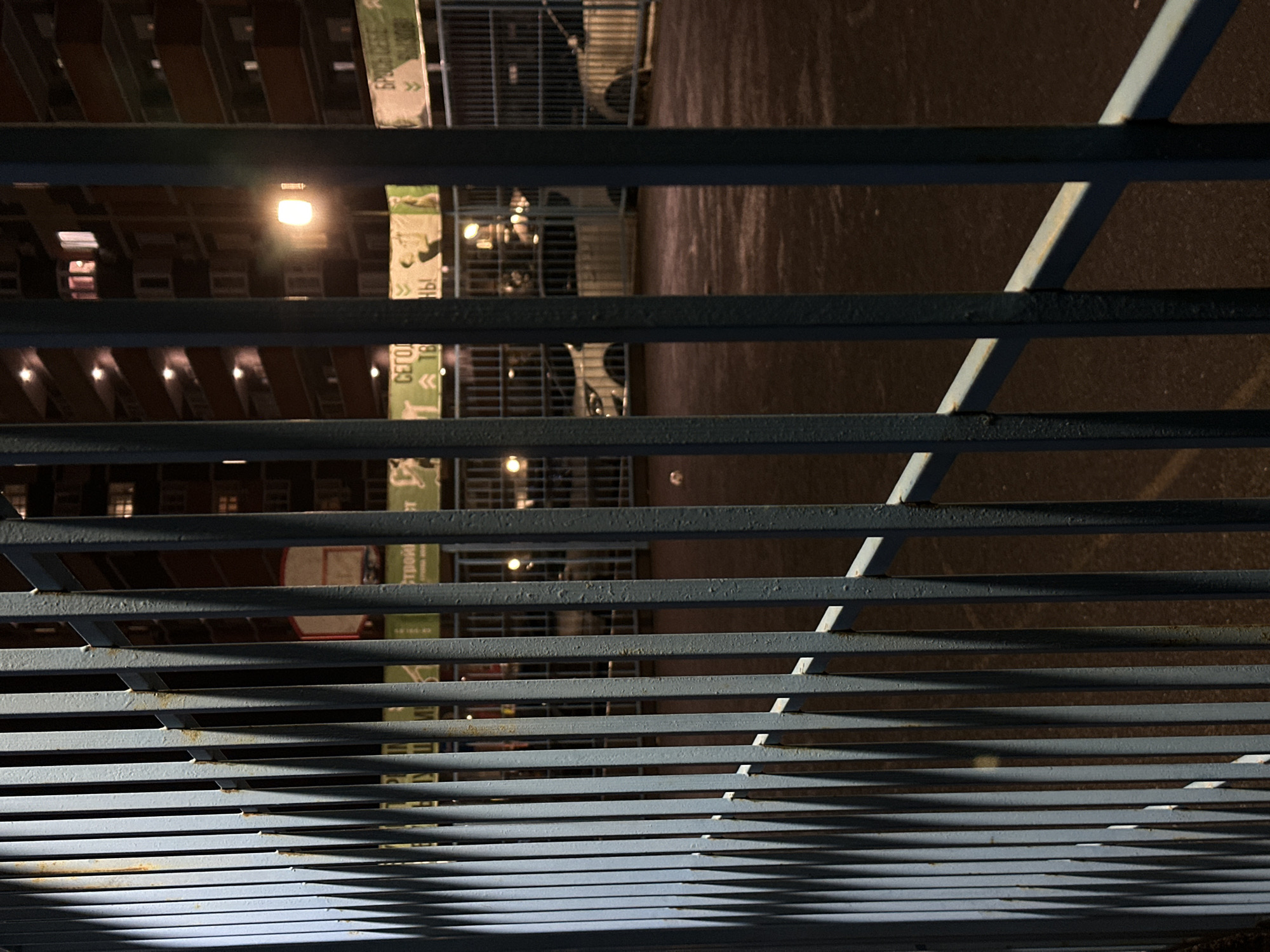 The image shows a metal fence in the city of Kuleshovka, Russia. The fence is gray-blue and runs horizontally across the frame. Behind the fence, a dark building with lit windows is visible. There is also a sign and some cars. The ground appears to be wet. The image seems to have been taken at night, given the darkness and artificial lighting.