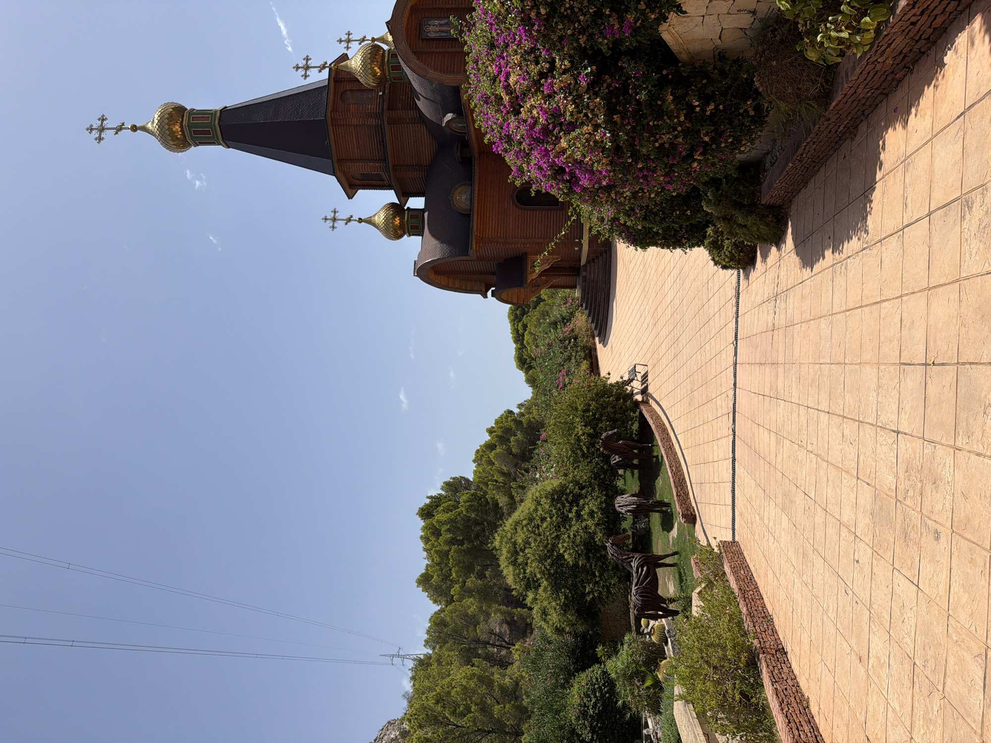 The image shows a bright, sunny day with a clear blue sky. In the foreground, a paved pathway with large square tiles leads towards a wooden building with golden domes and crosses, characteristic of an Orthodox church. The building is surrounded by lush green trees and vibrant purple bougainvillea flowers. To the left of the path, there are several metallic sculptures of horses on a grassy area. In the background, power lines stretch across the sky, and a rocky hillside covered in trees is visible. The scene appears to be located in Altea, Spain, likely a religious or cultural site. There are no people or animals visible in the image.