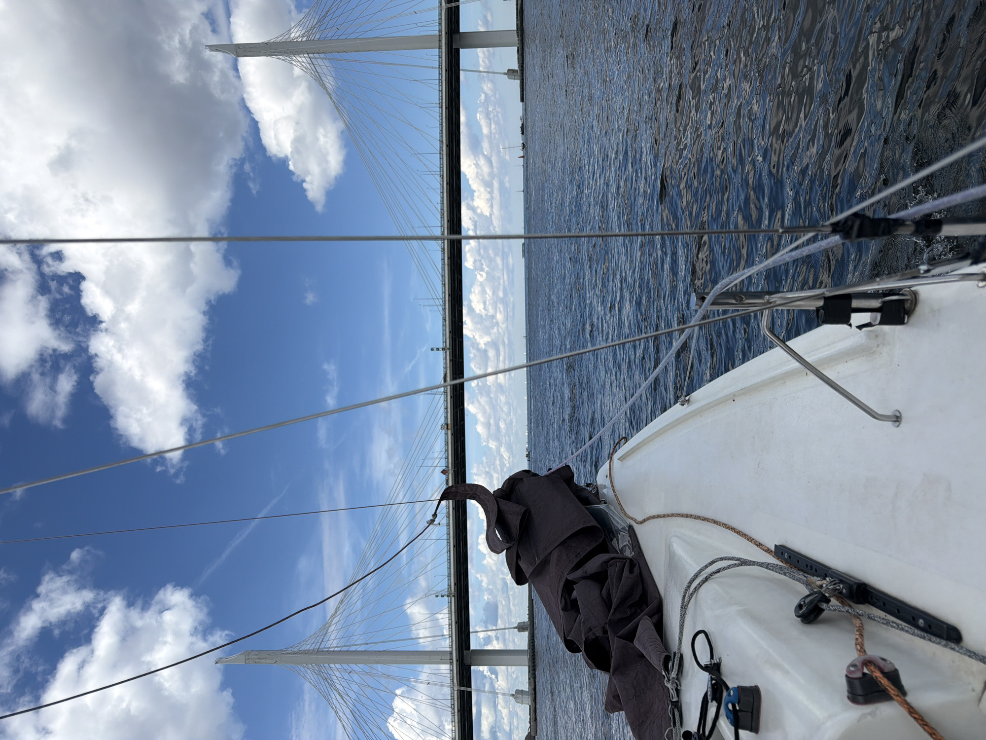 The image shows a view from a sailboat sailing on a body of water. In the background, a large cable-stayed bridge spans the water. The bridge has tall white pylons and a wide roadway with traffic. The sky is blue with scattered white clouds. There are other sailboats visible in the distance. The sailboat in the foreground is white and has a dark sail partially furled. The water is dark blue and choppy. The overall scene suggests a pleasant day for sailing near a major urban area. The bridge is the Western High-Speed Diameter in Saint Petersburg, Russia, a prominent landmark of the city.