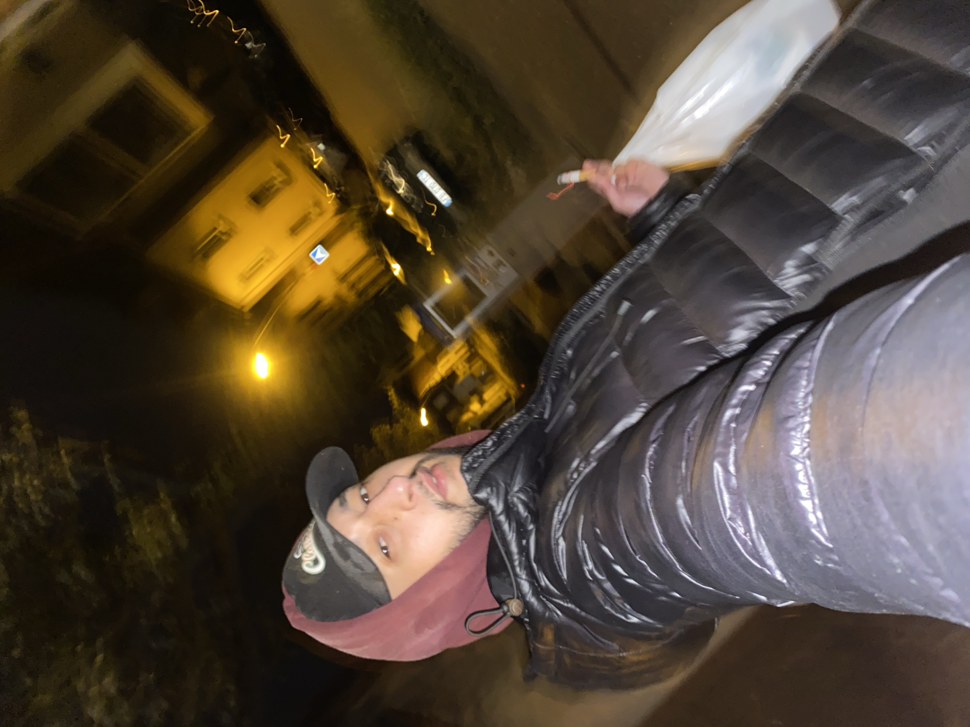 A man takes a selfie outside at night. He wears a black baseball cap, a maroon hoodie, and a black puffer jacket. In his hand, he holds a lit cigarette and a translucent white plastic bag. The background features illuminated buildings, blurred and out of focus, indicating a street scene in Schiltigheim, France. Street lighting casts a warm glow on the buildings and surrounding environment.