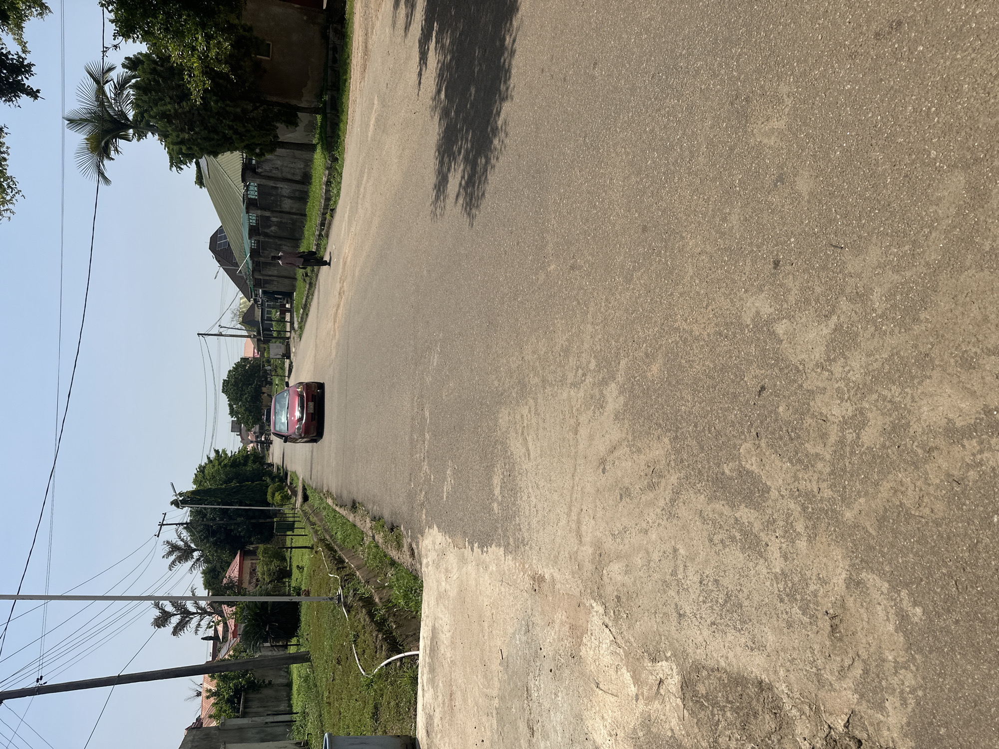 This image depicts a street scene in Abuja, Nigeria, likely in a residential area. The foreground shows a textured concrete surface merging into an asphalt road. Along the right side of the road, trees cast dappled shadows. A red car is stationary in the middle of the road, and a person is walking away from the camera on the right sidewalk. The left side of the road features a grassy area with a drainage ditch and utility poles supporting overhead wires. In the background, houses with red and green roofs are visible, interspersed with trees and foliage. The sky is clear and bright, suggesting daytime.