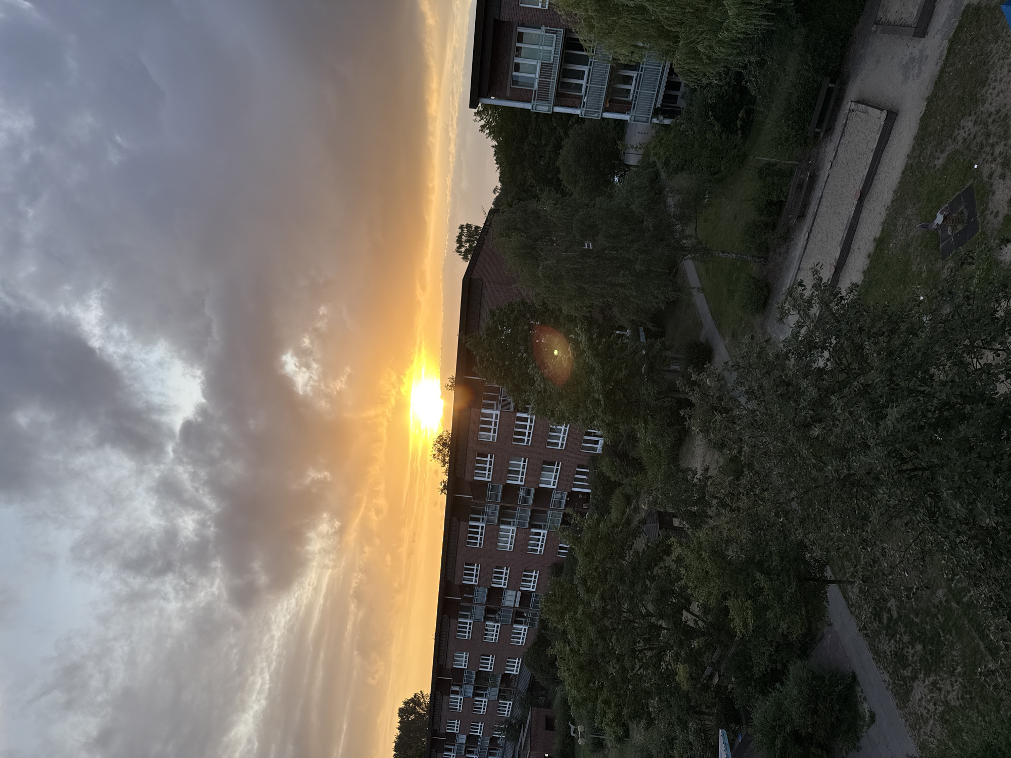 The image depicts a sunset over a residential area in Hamburg, Germany. The sky is filled with a dramatic mix of orange, yellow, and gray clouds, with the sun partially obscured by the clouds. In the foreground, there are two apartment buildings made of red brick with white-framed windows and balconies. Lush green trees and bushes surround the buildings, creating a sense of urban greenery. A playground with a sandpit and some play equipment is visible in the lower right portion of the image. The overall scene conveys a calm and serene evening atmosphere.