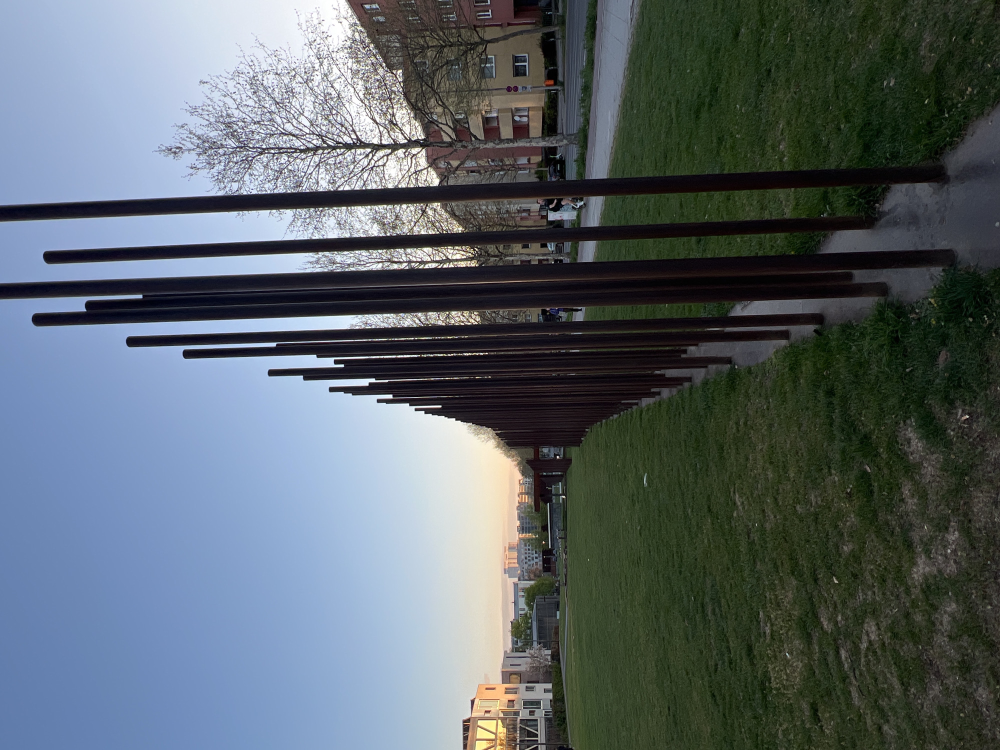 The image, when viewed upright, depicts a section of the Memorial to the Murdered Jews of Europe in Berlin, Germany, captured during the late afternoon or early evening. A prominent feature is the long array of dark, weathered, vertical concrete stelae (pillars) extending from the left foreground into the distance. These stelae vary in height and appear to line a paved path, which runs parallel to a large expanse of green grass on the right side of the frame.
The sky, occupying the upper left quadrant, is clear, transitioning from a vibrant blue overhead to a lighter, warmer, pale orange-pink hue near the horizon, indicative of a sunset or late daylight. A deciduous tree with bare branches is silhouetted against the bright sky in the upper left. In the mid-ground, beyond the memorial's structures, urban buildings, including multi-story residential blocks with varied facades, are visible along a street or walkway. A few indistinct figures of people can be seen moving between the stelae and the buildings. The far distance reveals a hazy cityscape against the soft, transitional light of the sky. The foreground features green grass with some visible dry patches. The scene blends the solemnity of the memorial with the surrounding urban environment under a tranquil sky. No visible text is present.