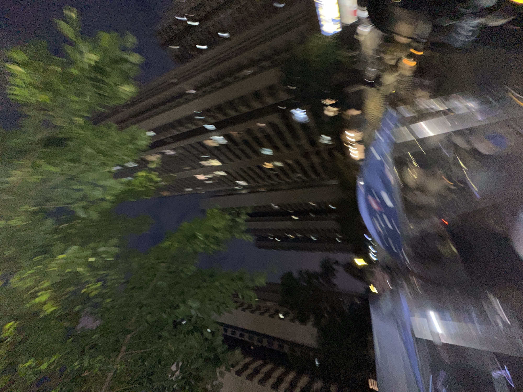The image depicts a nighttime scene in an urban environment. A tall building is visible in the background, with numerous illuminated windows. Trees with dense foliage are present in the foreground and partially obscure the view of the building. A shiny, reflective surface appears on the right side of the image, possibly part of a vehicle or building facade, reflecting light and distorting the surrounding environment. The location is presumably the Chongqing High-tech Zone in China, given the prompt's specification. However, this cannot be verified from the image alone.
