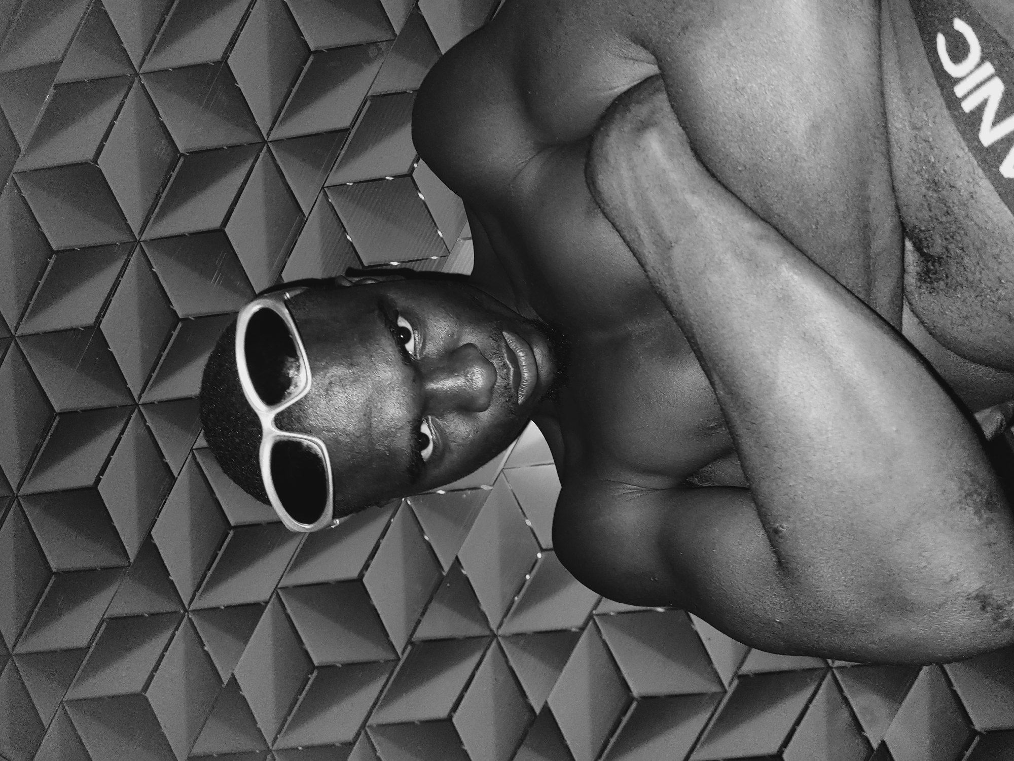 A black and white, high-angle, medium shot shows a person lying down on a textured surface. The person is wearing sunglasses with white frames and is positioned with their head facing the viewer. Their body is curled up, with their arms wrapped around their legs. The background is a tessellated pattern of dark gray rhombuses. The person appears to be male, with dark skin and short, dark hair. They are wearing a garment with the word "NICE" printed on it in white letters. The overall mood of the image is intimate and contemplative. The location of Bukuru, Nigeria is not depicted in the image.
