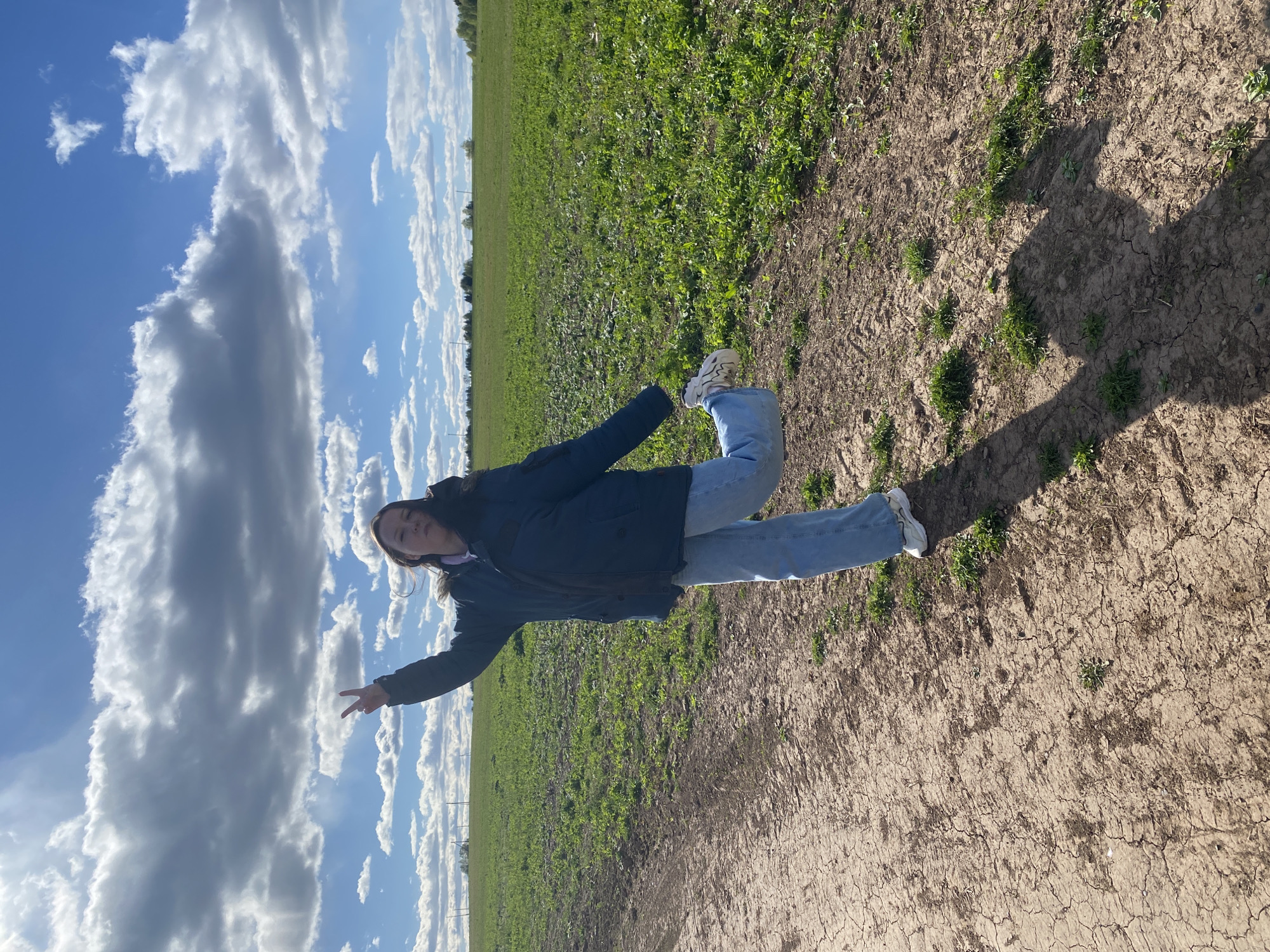 A female wearing a dark blue coat and light blue jeans is posing in a field in Дедушкино, Russia. She has her arms outstretched, making a peace sign with one hand. The sky is blue with cumulus clouds, and the ground is dry and cracked with patches of green grass. A shadow falls on the ground near her feet.