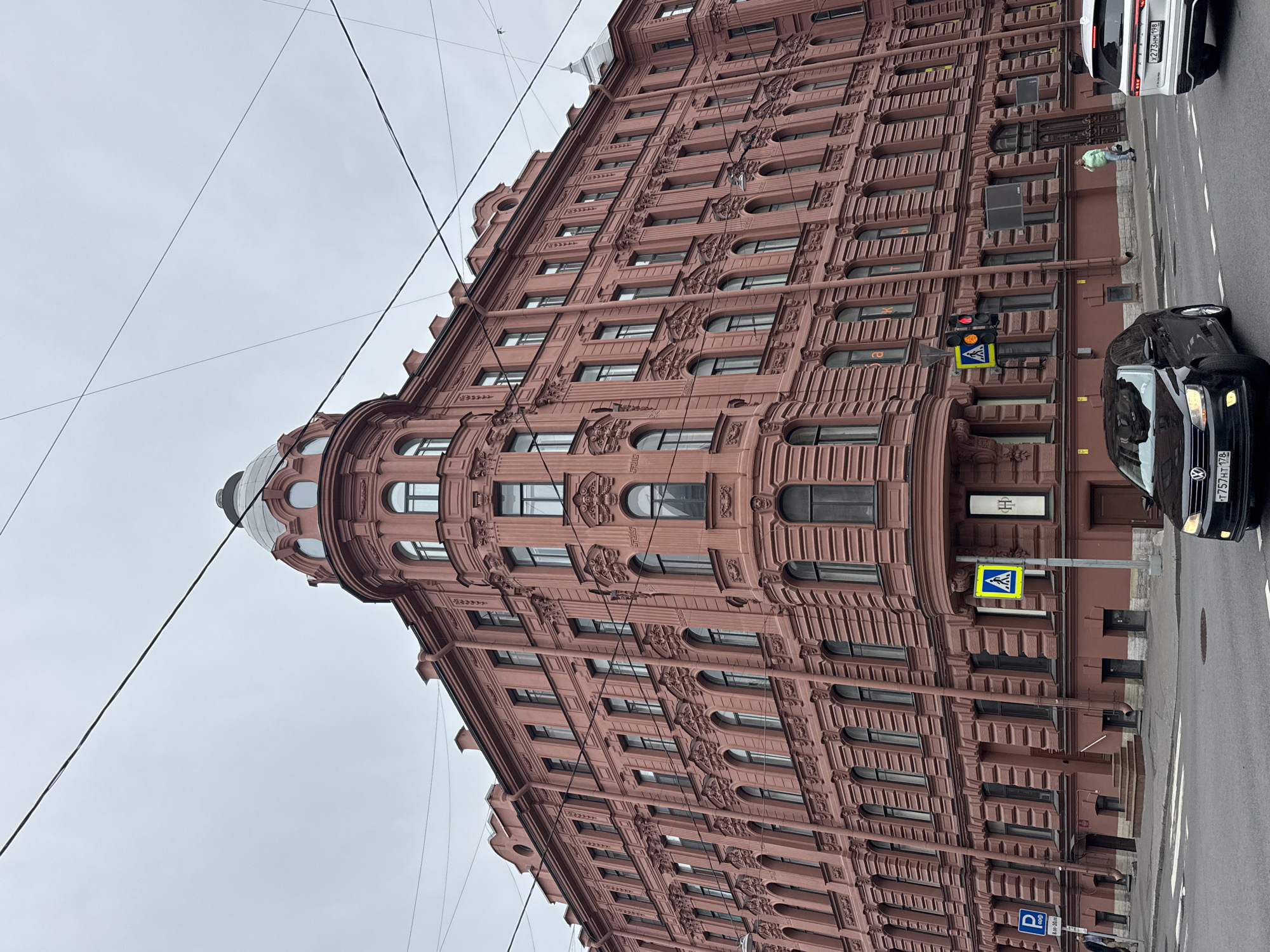 The image shows a street scene in Saint Petersburg, Russia. The foreground features a brown, ornate building with multiple floors and arched windows. Power lines are visible against a grey sky. A pedestrian crossing sign is placed next to a traffic light displaying red. A black car is driving on the road, and a white vehicle is partially visible in the upper right corner. A person is standing on the sidewalk near the building. A parking sign with a "P" is visible near the bottom.