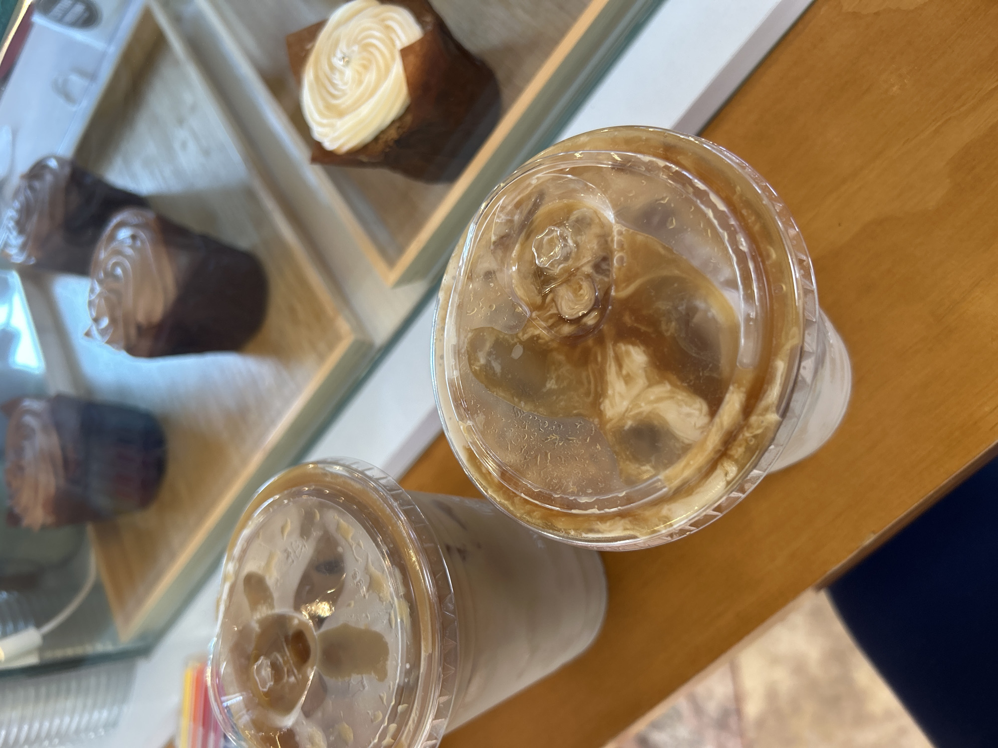 This image, taken from a slightly elevated, tilted perspective, captures a scene at what appears to be a cafe or bakery. The primary subjects are two iced beverages in clear plastic cups with domed lids, resting on a light brown wooden countertop. The drink in the foreground, closer to the upper right, shows a distinct swirl of cream or milk mixing with a darker beverage, likely coffee or tea, over ice cubes. The second drink, partially visible to its left and slightly behind, also contains ice and appears to be a creamy iced drink.
In the background, on the left side of the frame, is a glass display case containing several pastries. One prominent pastry, near the top left, is a cupcake or muffin topped with a neat swirl of white or cream-colored frosting, adorned with a small, shiny silver detail. Further back and to the left, several other dark-colored pastries, possibly chocolate muffins or cupcakes, are arranged on light-colored wooden or beige shelves within the display. The glass of the case reflects some ambient light.
The scene suggests a casual setting, possibly during a break or a visit to a coffee shop. There are no people visible, and no discernible text. The lighting appears soft and even, typical of an indoor environment.