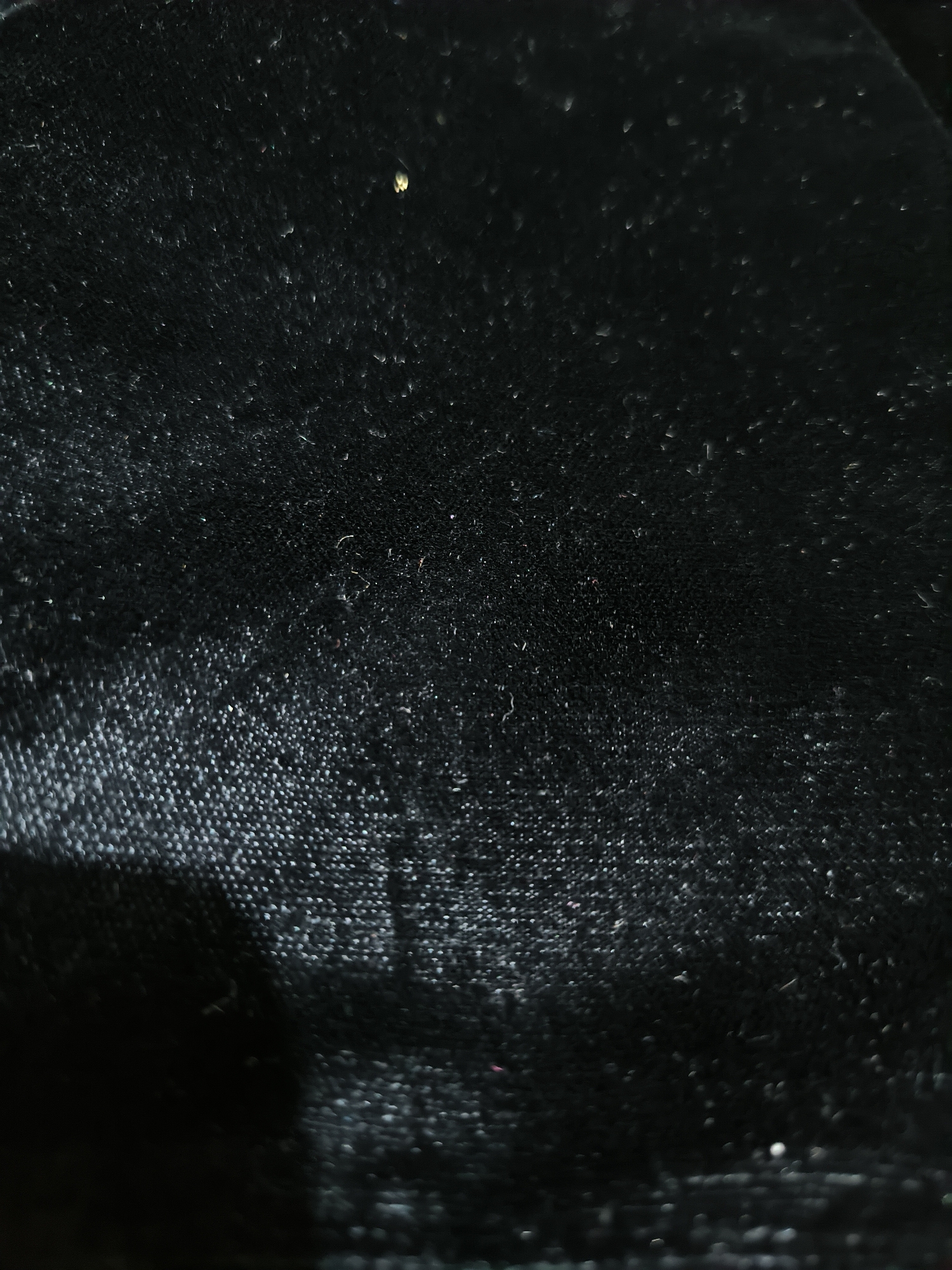 The image shows a close-up of a dark, textured surface, possibly fabric or a similar material. The lighting is dim, with variations in brightness across the surface, creating an effect of depth. Minute details like small fibers or particles are visible, enhancing the texture. The image lacks specific contextual information regarding location, people, objects or actions.