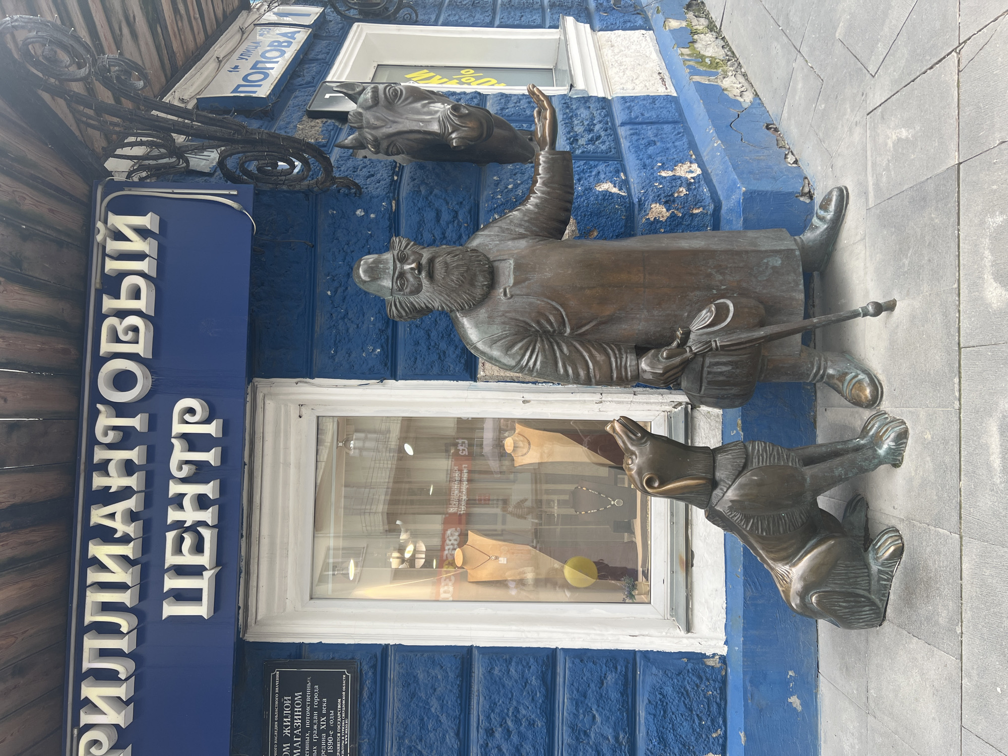The image depicts a bronze sculpture of a man and a dog in front of a building in Yekaterinburg, Russia. The building has a bright blue facade with white trim around the windows and signage. The signage above the window reads "БРИЛЛИАНТОВЫЙ ЦЕНТР" (Brilliant Center). To the right of the window, there is a street sign indicating "УЛ. ПОПОВА" (Popova Street).
The sculpture on the right shows a man with a long beard, wearing a hat and an apron, holding a horse's head in his outstretched left hand. He is also holding a walking stick with a bag attached to it in his right hand. The dog sculpture is on the left, sitting and looking up. The sculptures are positioned on a paved sidewalk in front of the building. The window displays jewelry on mannequins.