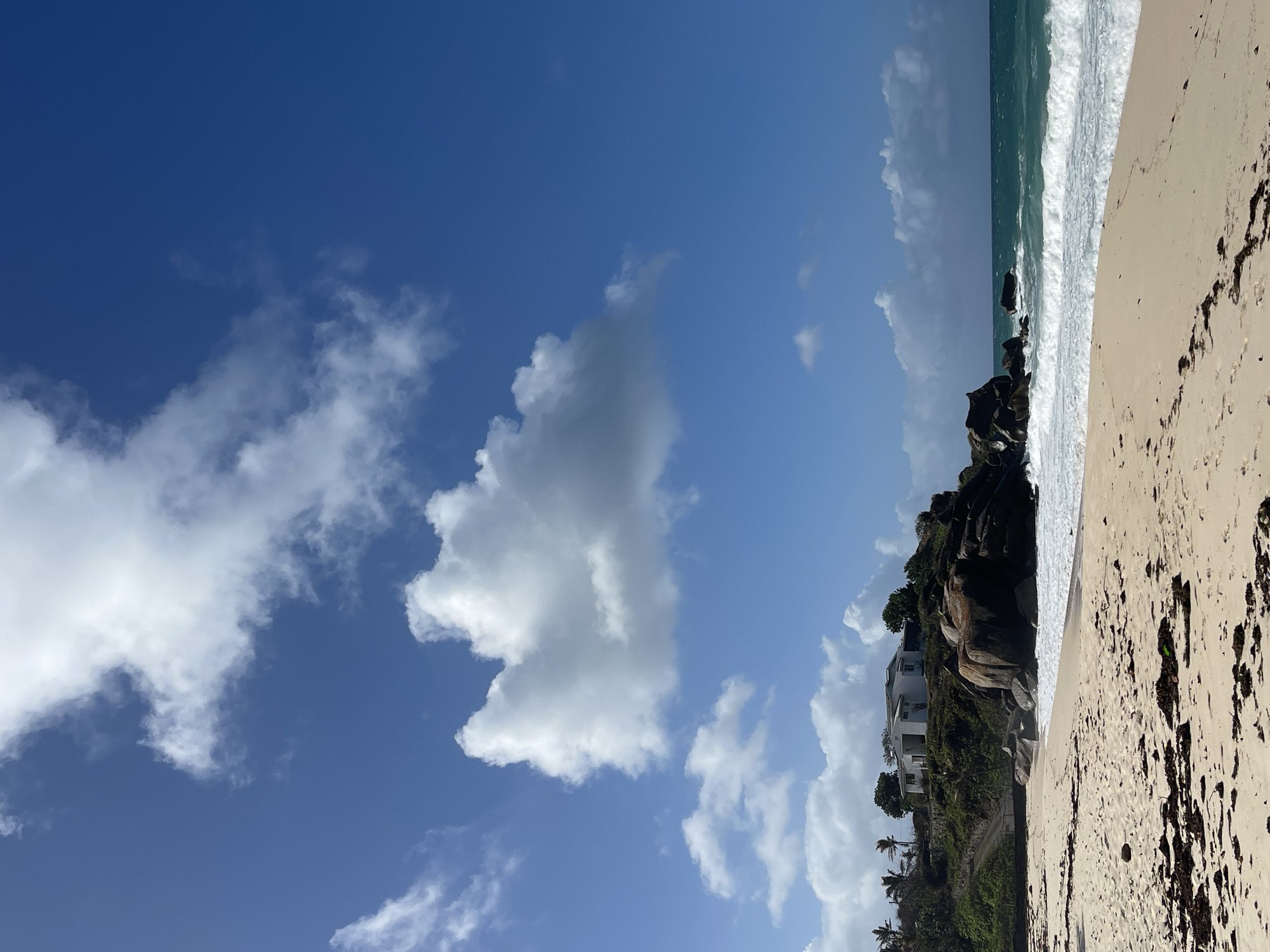 The image depicts a tranquil beach scene in Vista do Mar, Seychelles. The foreground is dominated by a sandy beach with scattered dried seaweed and footprints. In the midground, the turquoise ocean meets the shore with gentle waves. To the right, large, dark rocks jut out into the water. Perched on a verdant hill overlooking the ocean, a white house with a light-colored roof stands prominently. The house is surrounded by lush green trees and vegetation. The sky above is a vibrant blue, dotted with fluffy white clouds, indicating fair weather. The overall scene conveys a sense of peaceful isolation and natural beauty characteristic of the Seychelles.