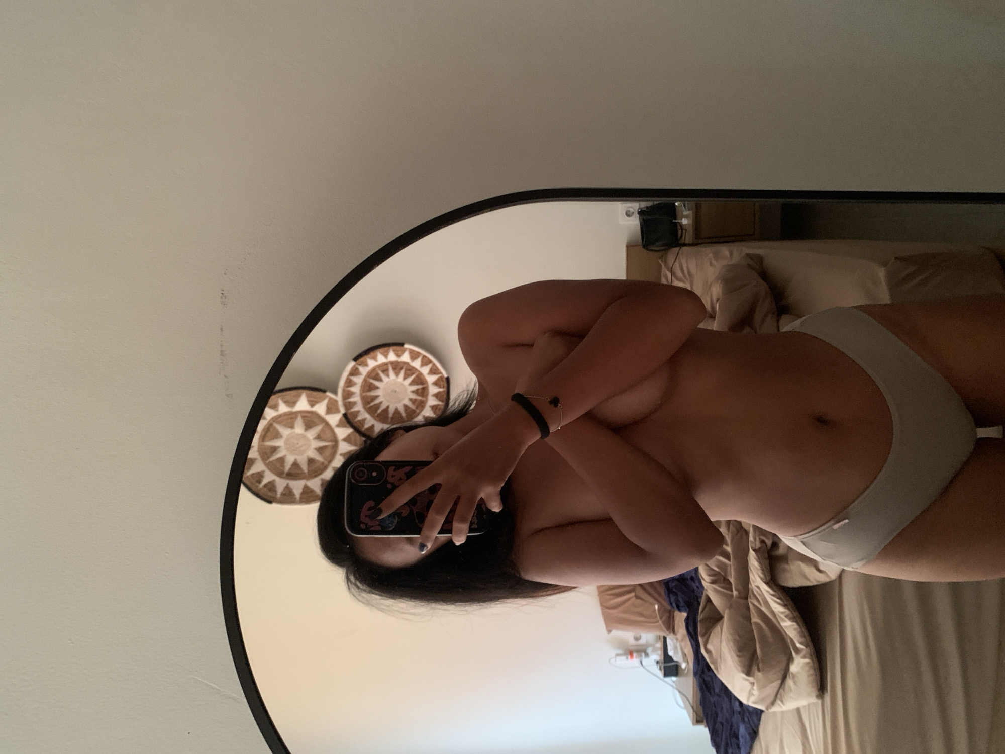 A person is reflected in a round mirror, holding a smartphone with a black patterned case in front of their face. They are wearing light gray underwear and are covering their chest with their arms. The person has dark hair. Behind the person, in the background, there is a bed with tan-colored bedding, including pillows and a duvet. Additionally, there is a dark blue blanket partially covering the bed. On the wall, adjacent to the mirror, there are three decorative circular wall hangings. The specific location in Kuta Selatan, Indonesia is not discernible from the image.