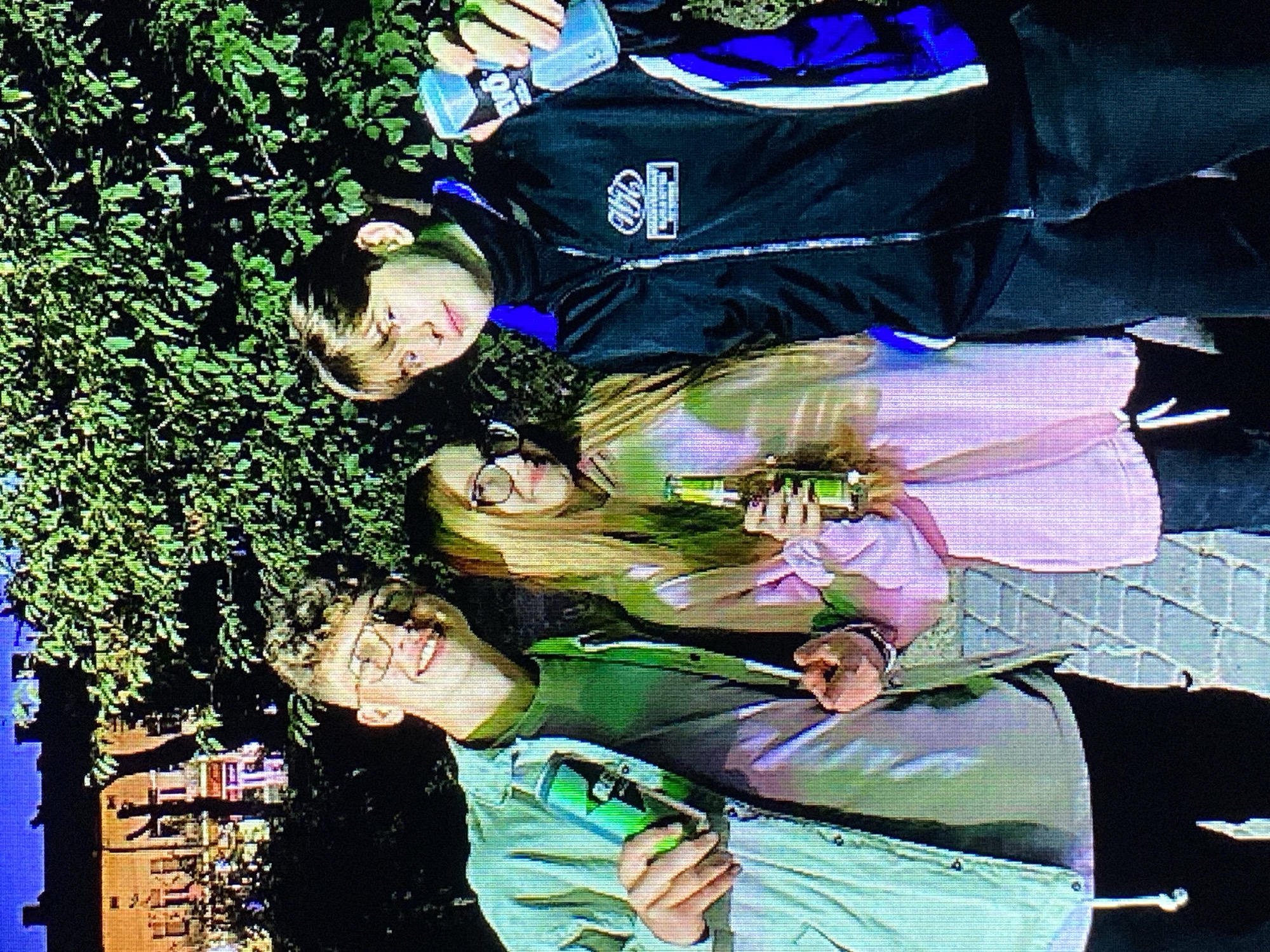 Three young people are standing close together outdoors at night, in front of lush green foliage and a building in the background. On the left, a male with curly hair and glasses is holding a green can. He is wearing a light blue jacket over a green t-shirt and black pants. In the center, a female with blonde hair and glasses is holding a brown bottle and wearing a pink hooded sweatshirt. On the right, a male with light brown hair is holding a blue and silver can. He is wearing a black jacket with blue accents over a black turtleneck and dark pants. The building in the background appears to be in a European style, with illuminated windows and architectural details. The lighting suggests an urban setting, possibly a street or park area in the city of Poznan, Poland. The pavement in the foreground has a patterned design.