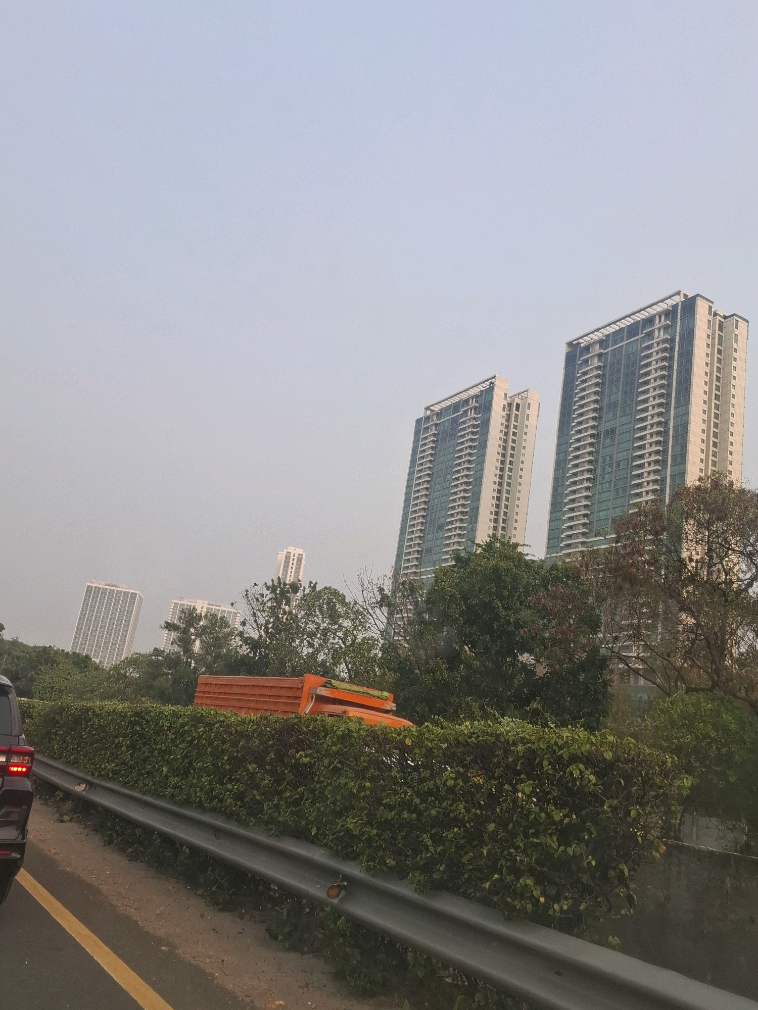 The image depicts a view of several modern high-rise buildings in the city of Panunggangan Barat, Indonesia. In the foreground, a black vehicle is partially visible on the left side of the road. A yellow line marks the edge of the road. A grey metal guardrail runs along the side of the road, separating it from a dense green hedge. Behind the hedge, the tops of trees and an orange truck are visible. The sky is overcast and hazy. The architecture of the buildings suggests a contemporary urban setting.
