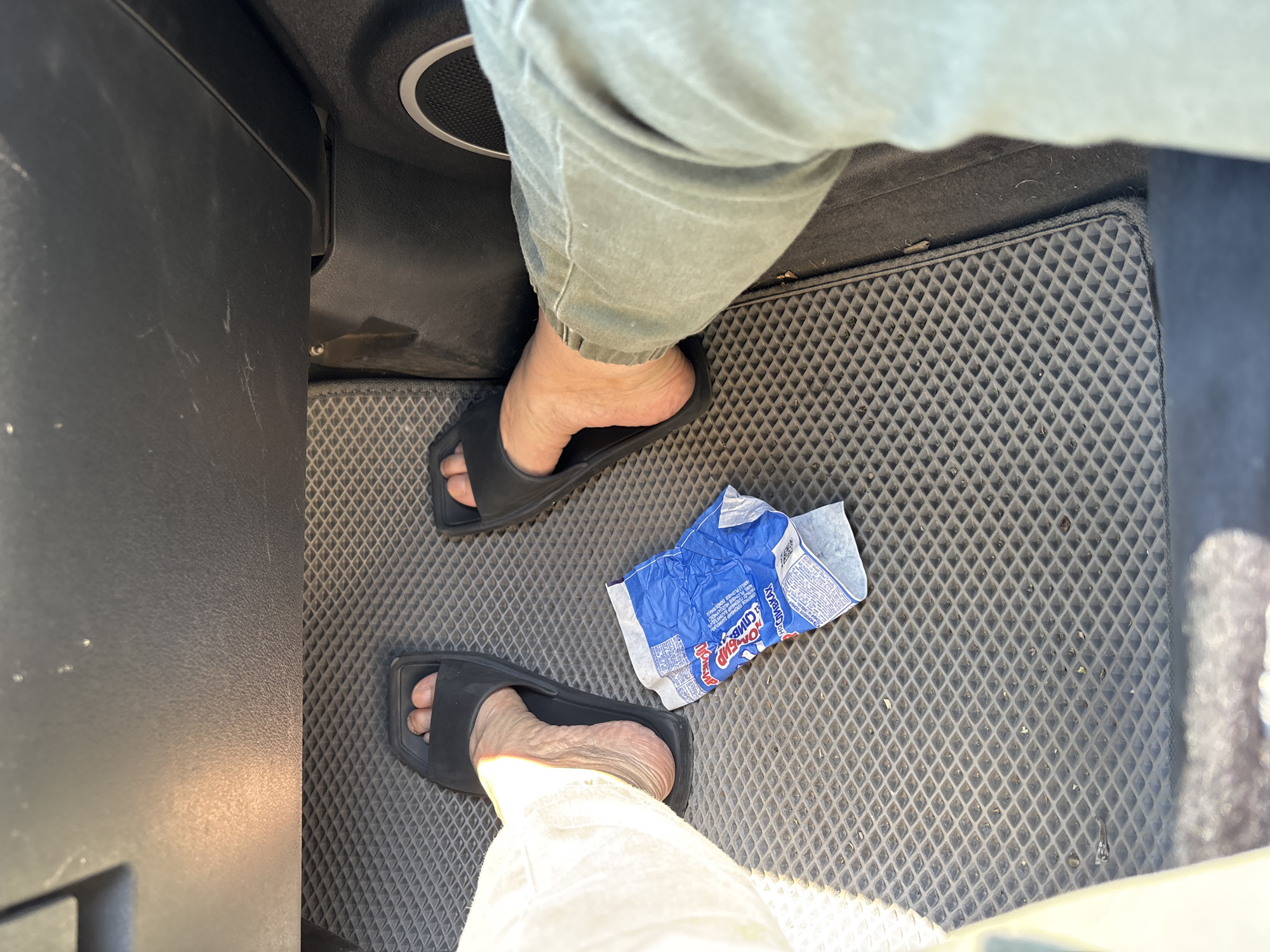 The image shows the feet of a person wearing black slides, resting on a gray textured car mat. A crumpled blue and white wrapper lies on the mat between the feet. The person is wearing light-colored pants on their left leg and olive-green pants on their right leg. The car's interior is visible above the feet, with a dark panel and a circular speaker grill. The scene is a car interior, likely in the city of Borodino, Russia.