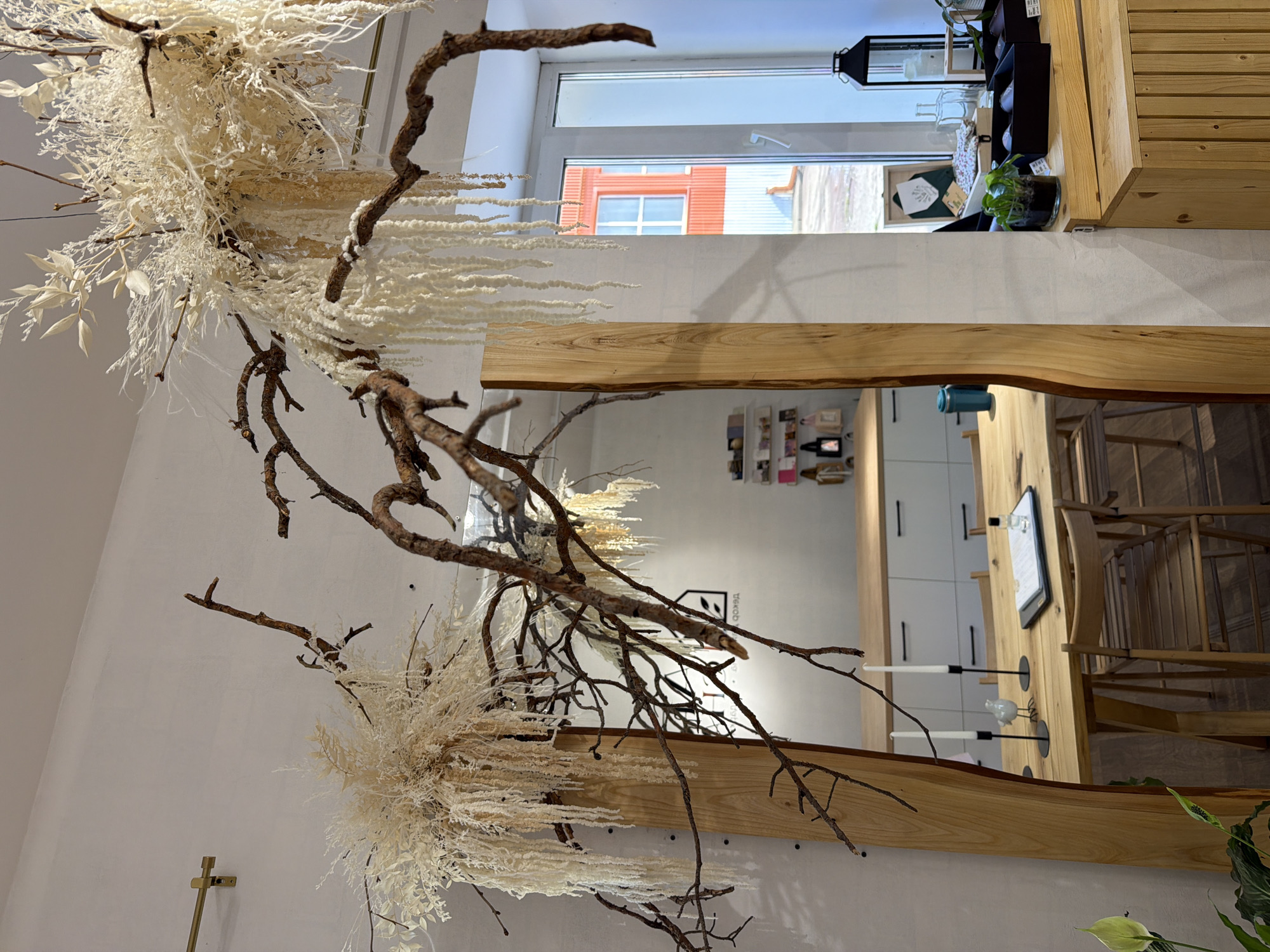 The image depicts the interior of a retail establishment, possibly a flower shop or a home decor store, located in Chaikovsky, Russia. The scene is dominated by a large, natural wood-framed mirror that reflects a wooden table set with two white taper candles in black holders, a small white ceramic bird figurine, and a clipboard with a document and a small glass bottle. Wooden chairs surround the table. Above the table and mirror, dried floral arrangements and branches hang from the ceiling, adding a bohemian and natural aesthetic to the space. In the background, a window reveals an orange building with white window frames, suggesting an urban or town setting. Shelving units are visible, displaying various items, including small cards and bags. The overall ambiance is rustic and minimalist. No people are visible in the image.