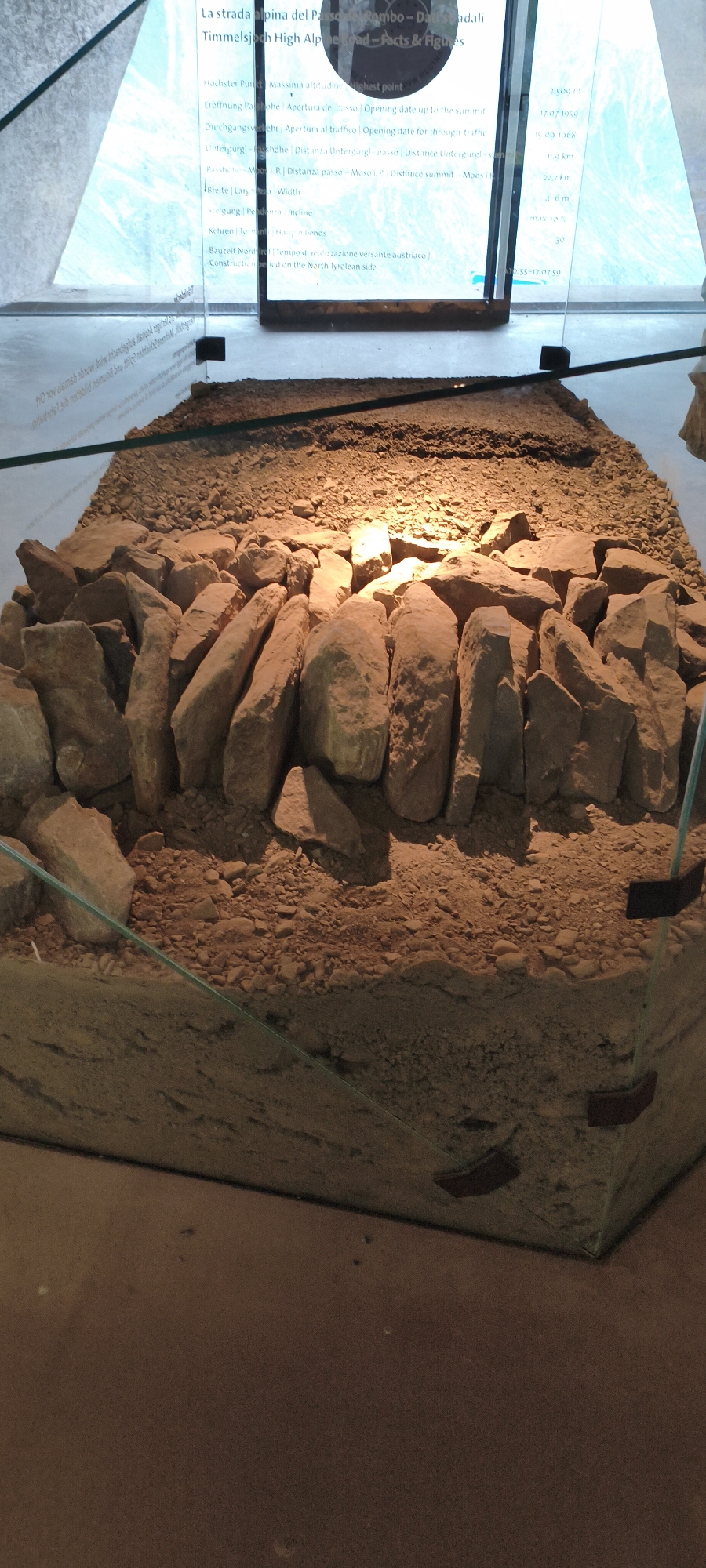 The image shows an exhibit within the Sölden, Austria, ice Q museum, focused on the Timmelsjoch High Alpine Road. The foreground features a cross-section of the road's construction, displaying layers of gravel and large rocks, encased in glass. Above the exhibit, a digital display provides information about the road, including facts and figures, with text in multiple languages. The background is illuminated, suggesting an indoor setting with artificial lighting, and hints at a scenic view through a large window. No people are visible in the frame.