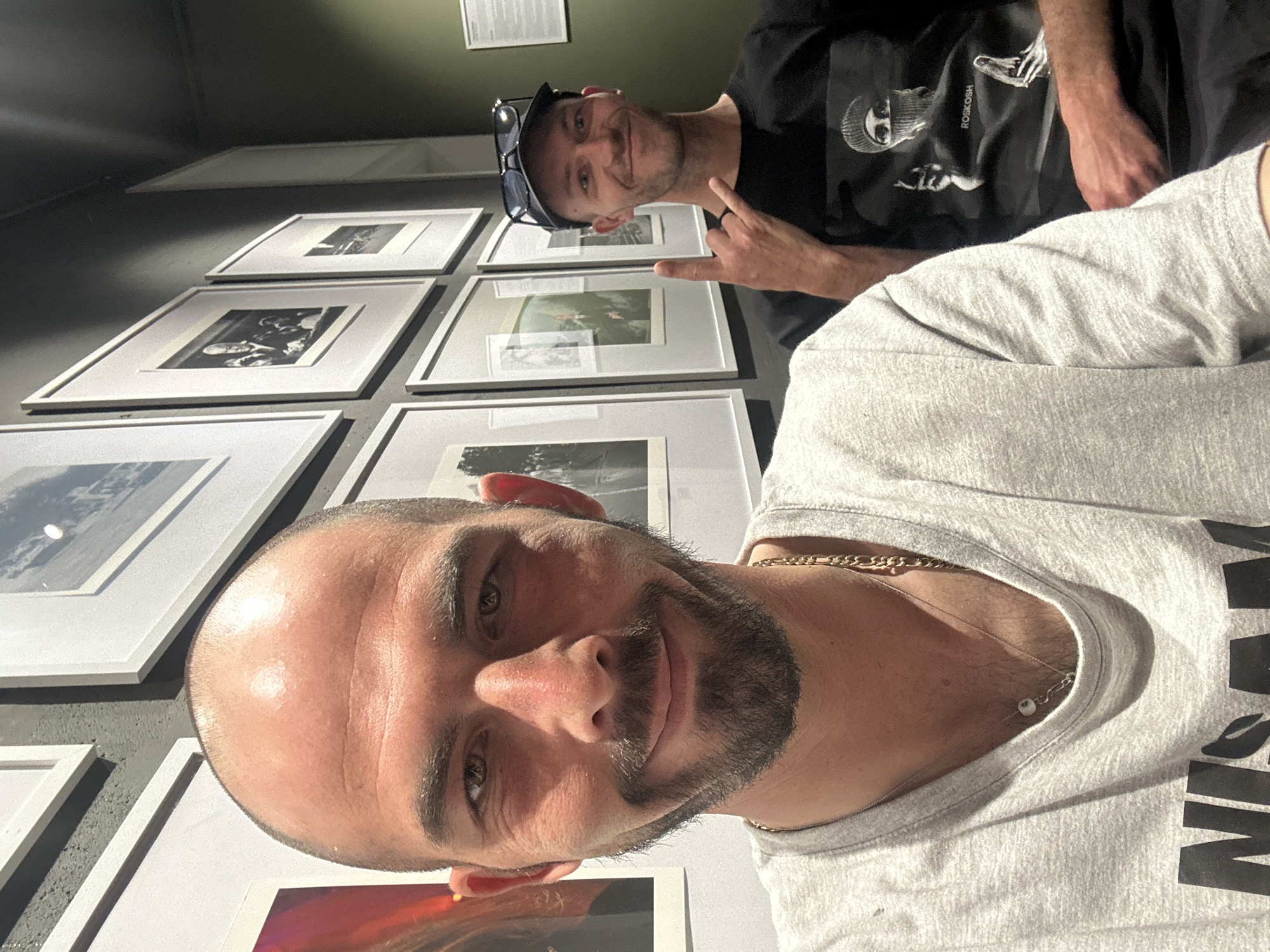 Two Caucasian males are posing for a selfie inside what appears to be an art gallery. The man in the foreground, closer to the camera, is bald with a trimmed beard and is wearing a light grey T-shirt with black lettering on it, and a gold chain and necklace. The other man in the background is wearing a black T-shirt with an image on it. He has sunglasses pushed up on his forehead and is giving a “rock on” hand sign. A wall of framed photographs is behind them. The exact location in Budva, Montenegro, is impossible to determine from the image.