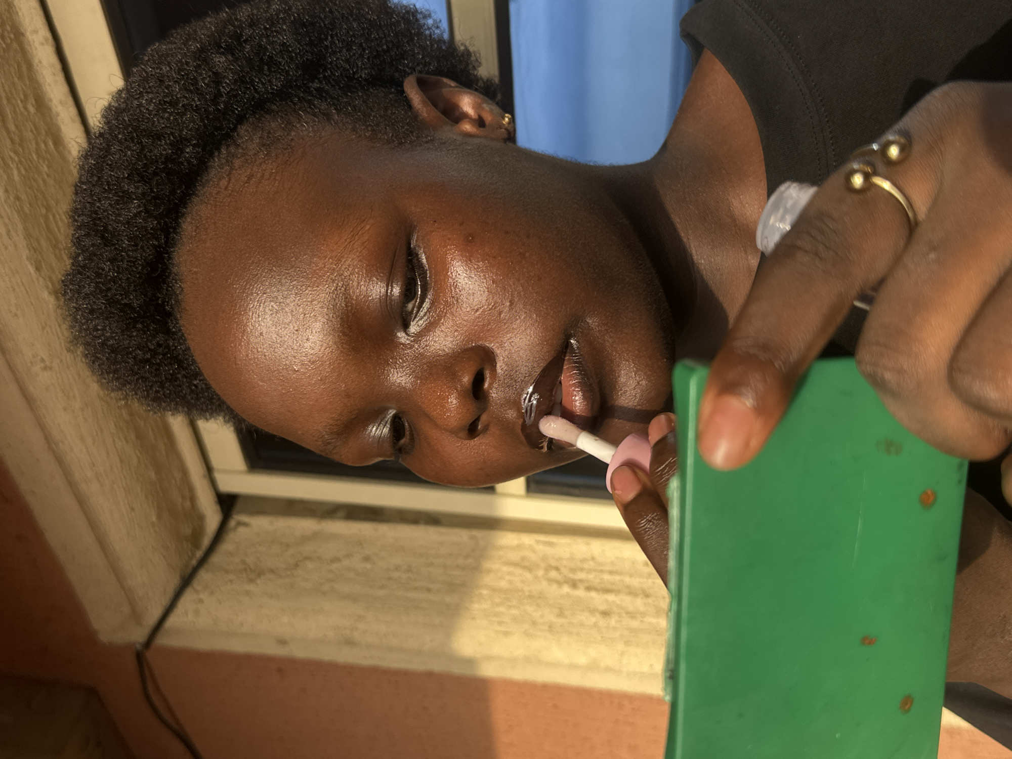 A young, dark-skinned person is applying lip gloss while looking in a green hand mirror. They are wearing a black top and a gold ring with two small spheres on their finger. The person is positioned near a window. The image was taken in Onitsha, Nigeria.