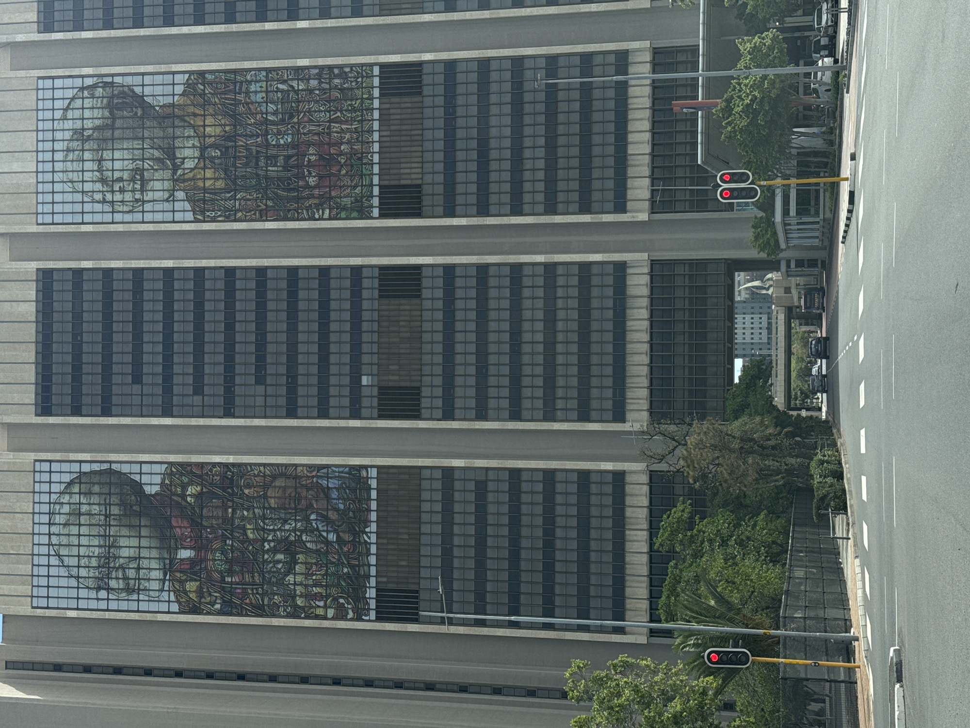The image shows a tall building in Cape Town, South Africa, with murals displayed on the building's facade. The murals depict a person's face and a complex decorative pattern. The building has a grey exterior with a grid-like window design. A street runs alongside the building, with a traffic light showing red. There are trees and other greenery visible along the street and in the background. The image suggests an urban environment with a combination of modern architecture and natural elements.