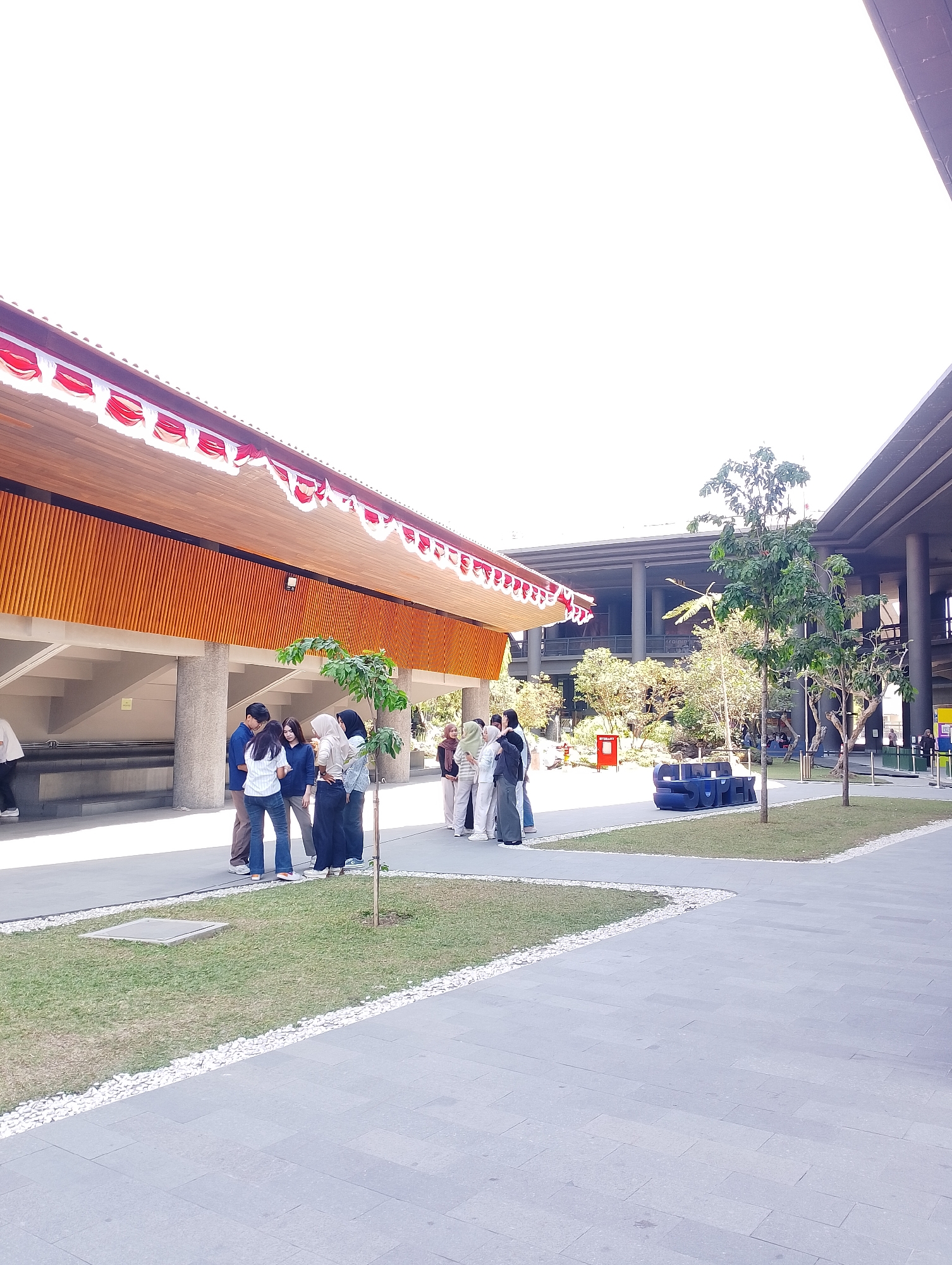 In Depok, Indonesia, a group of young adults are gathered outside a modern building with a distinctive overhanging roof decorated with red and white bunting. The building appears to be a public space or educational institution, possibly a campus, given the presence of lush greenery and paved walkways. The architectural style is contemporary, featuring large structural columns and multiple levels visible in the background. A blue sculptural element spelling out "SUPER" is situated on a grassy area, serving as a focal point. The individuals, some wearing hijabs, are dressed in casual attire, and their interactions suggest a relaxed, social atmosphere. The lighting indicates it is daytime, with bright, natural light illuminating the scene.