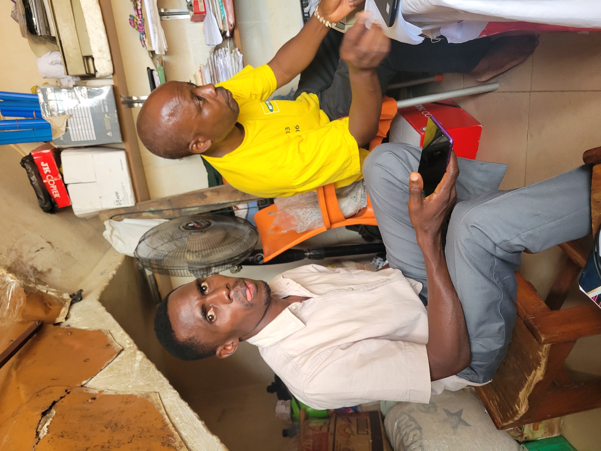 Two men are seated in an indoor, cluttered environment. The man on the left, wearing a light-colored collared shirt and grey pants, is seated on a wooden stool and holding a dark-colored smartphone. He is looking towards the camera with his tongue sticking out slightly. The man on the right, wearing a bright yellow t-shirt with a logo and black pants, is seated on an orange chair. He is looking down and appears to be interacting with papers and a laptop on a white-covered table in front of him. Various items are visible in the background, including boxes, shelves with papers, a fan, and a broken wall structure. The lighting is dim and appears to be artificial.