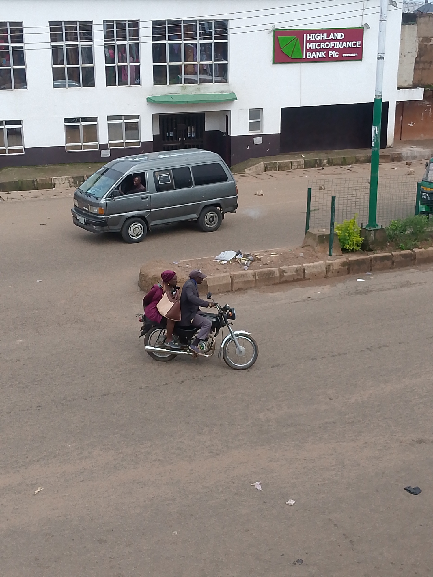 In Bukuru, Nigeria, a grey minivan drives down a paved street, its driver visible through the windshield. On the same street, a man and a woman ride a motorcycle. The man drives, wearing a cap and dark jacket, while the woman sits behind him, dressed in a purple outfit and carrying a tan bag. They are on the right side of the road, moving away from the camera. In the background, a white building with multiple windows and a sign for "HIGHLAND MICROFINANCE BANK PIC" is visible. The street is lined with a curb and some scattered debris. The overall scene depicts a typical urban environment in Nigeria.