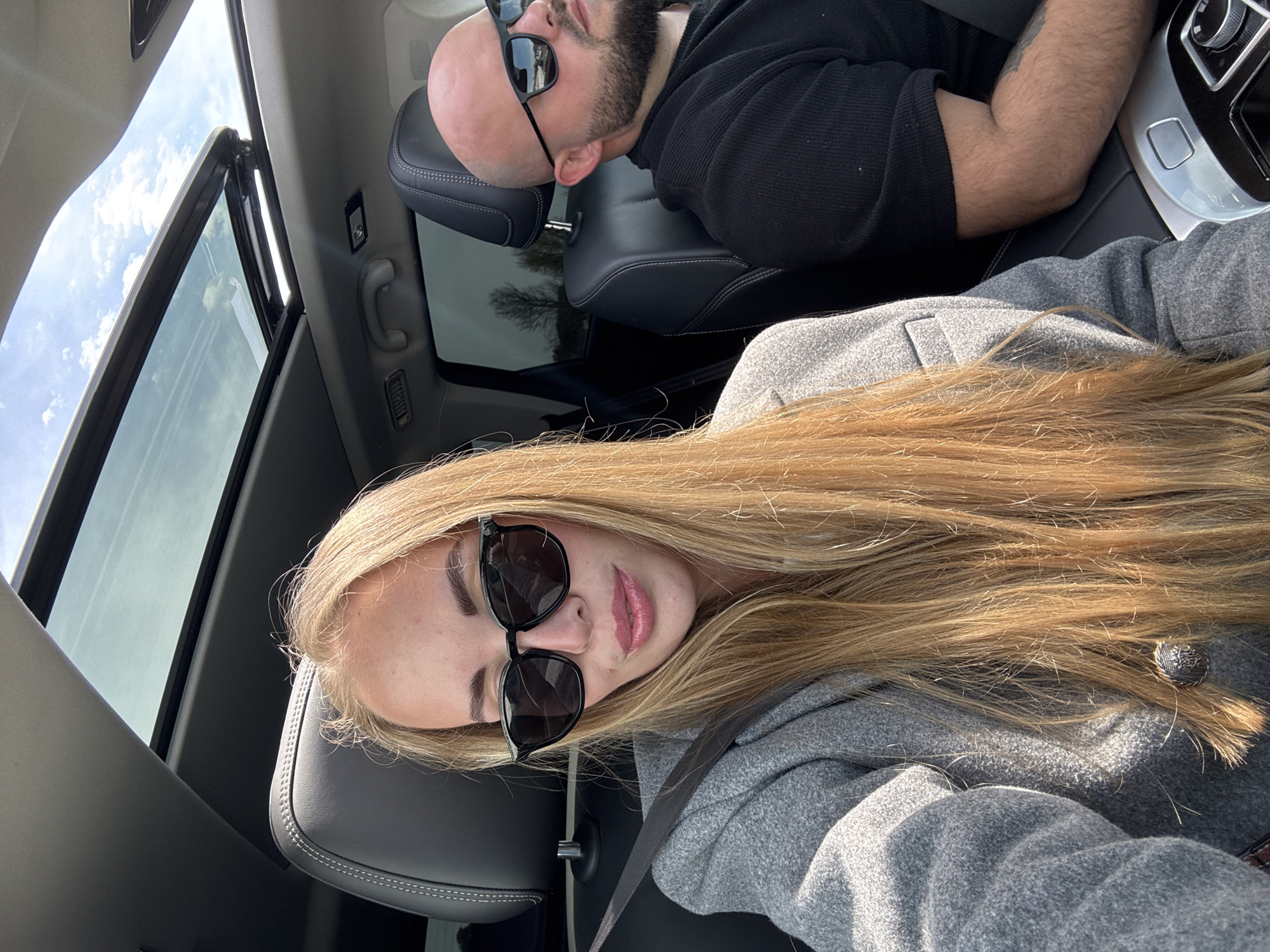 Two people are in a car with an open sunroof. The person in the foreground has long blonde hair and is wearing a grey jacket and black sunglasses. They are sitting in the driver's seat and wearing a seatbelt. The person in the background is bald, has a beard, and is wearing a black shirt and sunglasses. They are resting their head on the headrest. The interior of the car is primarily black leather and chrome. The location is not possible to be determined.