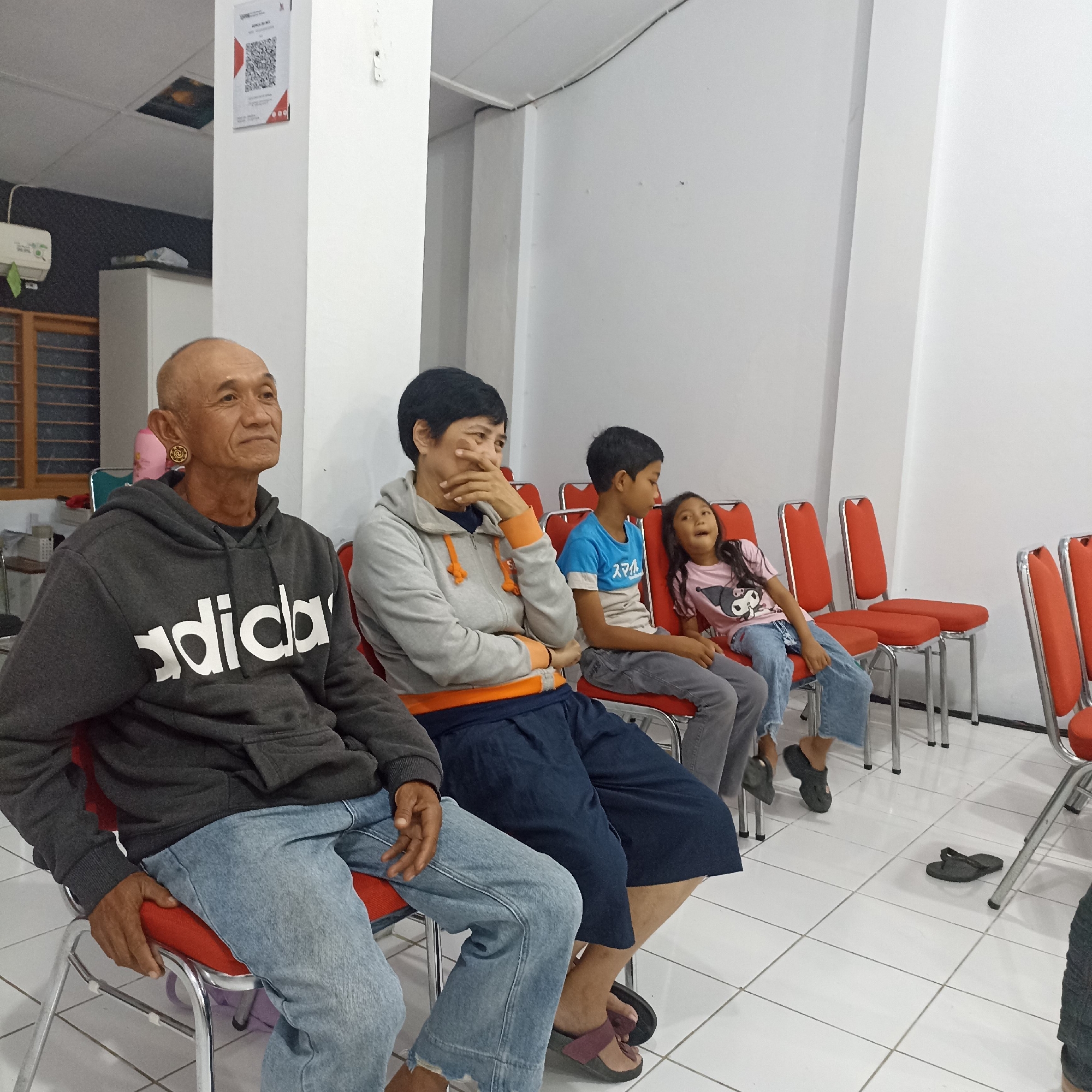 The image depicts an indoor scene with several individuals seated on red chairs. In the foreground, an older man with a shaved head is wearing a grey hooded sweatshirt with "adidas" printed on it in white letters, and blue jeans. He has a large, ornate earring in his left ear. To his right, a woman with short black hair is wearing a grey hoodie with orange accents and dark blue wide-leg pants. She is touching her face with her hand. Behind them, a boy in a blue t-shirt and grey pants is seated, looking to his right. Next to him, a young girl with long dark hair wears a pink t-shirt with a cartoon character and blue jeans. She is looking upwards with her mouth open. The room has white walls and a white tiled floor. A white pillar is visible on the left side of the image, with a QR code displayed on it. There are several red chairs lined up, with metal legs and frames. The overall environment suggests a waiting area or a casual gathering. There are no visible indicators that the location is in Magelang, Indonesia.