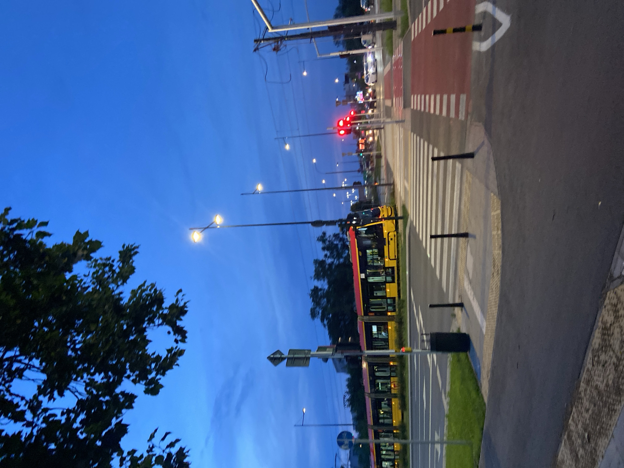 The image depicts a street scene at dusk in Warsaw, Poland. A yellow and red tram is stopped at a crosswalk, indicated by white stripes on the road. The tram has multiple windows and appears to be occupied by passengers, though their faces are not discernible. Traffic lights are visible and show red, suggesting a stop for vehicles. Several streetlights are illuminated, casting a soft glow. The sky is a deep blue, transitioning from twilight to night. Trees line the background, and power lines are strung overhead. The foreground shows a paved road and sidewalk. The overall impression is one of an urban environment at the end of the day.