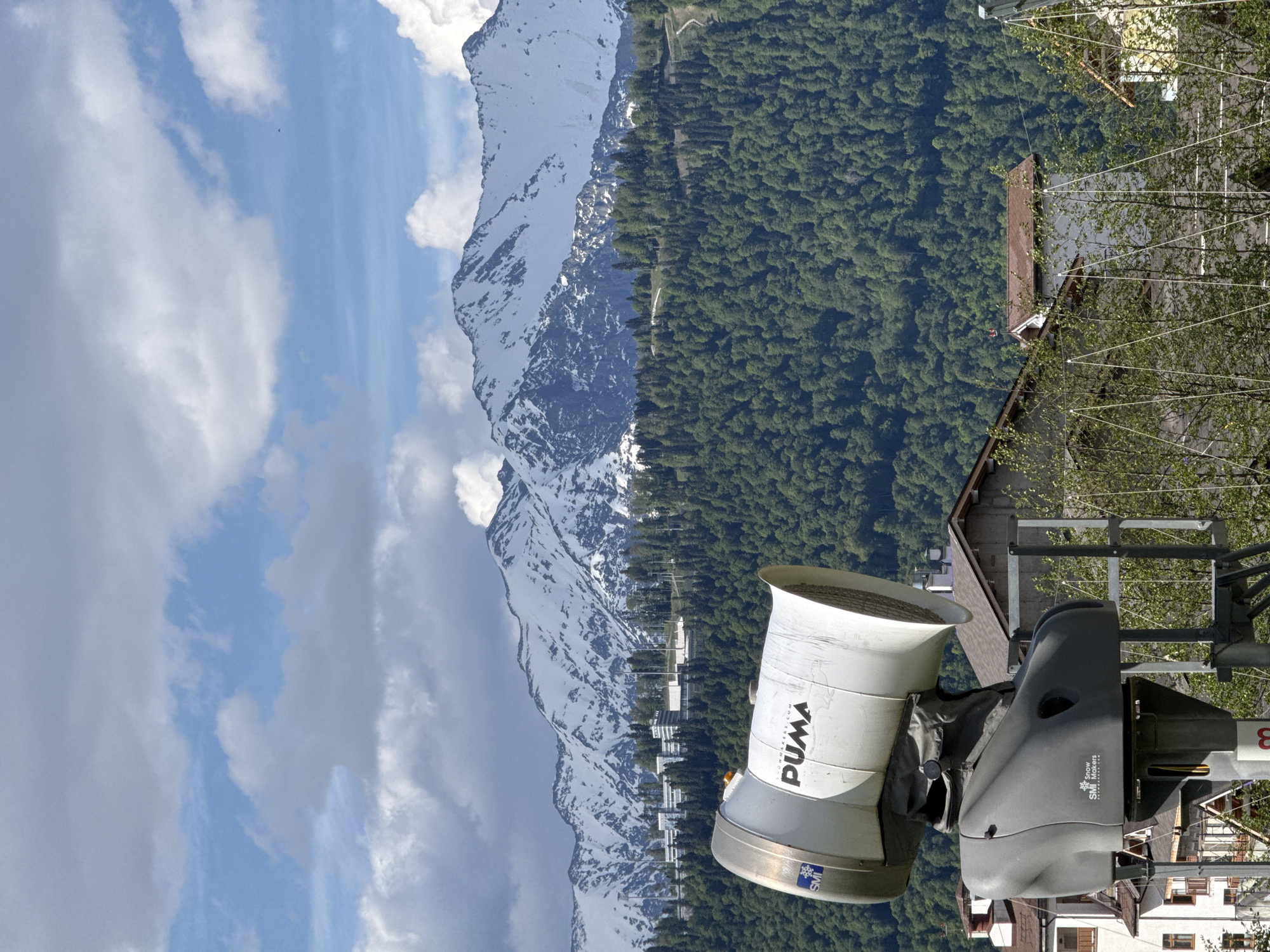 The image captures a vast mountainous landscape, likely a ski resort area in Estosadok, Russia, seen on a bright, clear day. In the foreground, on the right, a large white and grey snowmaking machine is prominently featured. Visible text on the snow cannon includes "PUMA", "TECHNICOM", and "SMI Snow Makers", along with an "SMI" logo.
The midground reveals a dense, green coniferous forest covering the lower and mid-slopes of a mountain. Faint lines suggesting ski lifts or cable car cables ascend through the trees, hinting at the area's primary use. Further into the distance, grand snow-capped mountains dominate the background, their peaks and upper reaches still covered in white snow, contrasting with the dark rock.
The sky above is a vibrant blue, adorned with soft, white cumulus clouds. A tiny dark speck, possibly a bird or a distant aircraft, is visible in the upper left portion of the sky. The time of day is clearly daytime, marked by strong sunlight, and the weather appears pleasant and clear. The combination of snow on the peaks and lush green foliage on lower elevations suggests a late spring or early summer season in this alpine environment, with the snow cannon currently inactive. The scene conveys the natural beauty and grandeur of a mountain resort.