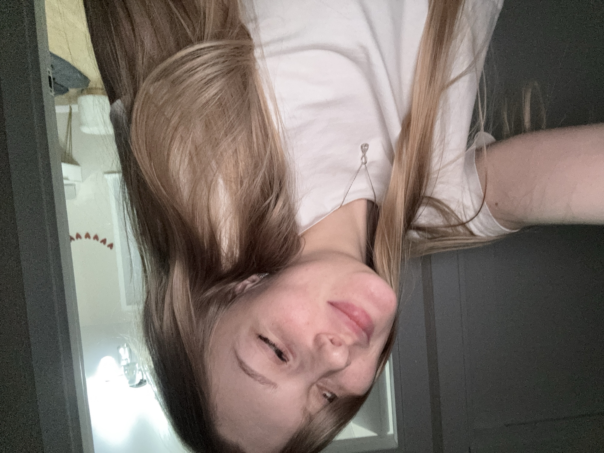 An upward-facing, rotated selfie captures a young woman from the chest up in an indoor setting, likely in Perm, Russia. Her long, light brown hair, with blonde highlights, falls around her face and shoulders. She wears a plain white t-shirt, and a delicate silver earring or piece of jewelry dangles near her collarbone on the right side.
The woman's head is tilted slightly, and she looks towards the camera with one eye partially closed, giving her a playful or squinting expression, possibly due to a bright light source. Her lips are slightly parted in a subtle smile or smirk.
In the background, visible behind her, are dark grey or black walls or doorways, suggesting an interior space. A bright, white light fixture is visible on the wall behind her. Further back, above the light, faint red, inverted V-shaped decorations are arranged in a row. What appears to be a broom or mop handle is also faintly visible in the background. The lighting suggests an indoor setting, possibly with artificial light. No visible text is present in the image.