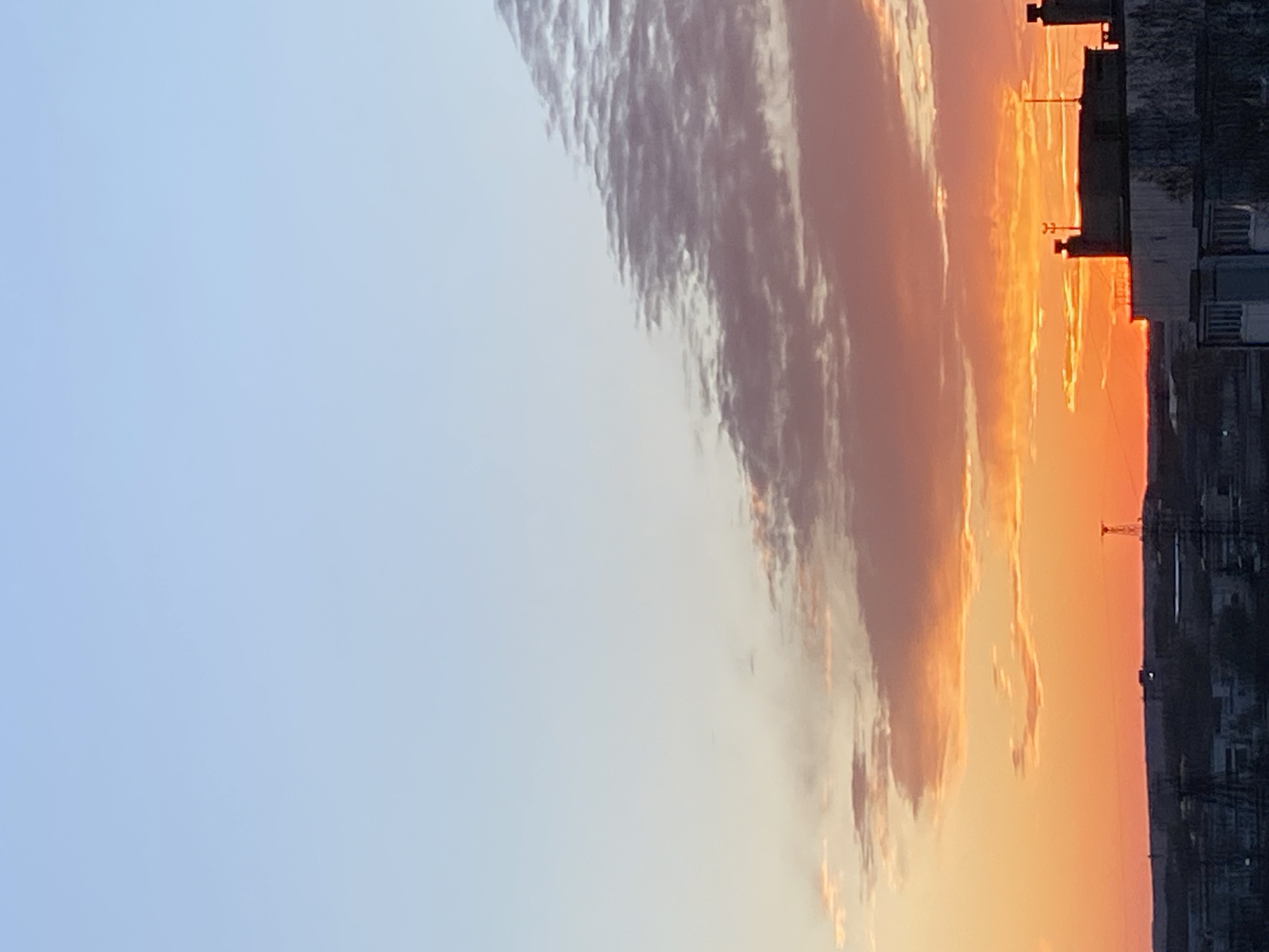 The image depicts a sunset in Chaikovsky, Russia. To the right, a silhouette of buildings and a communications tower are visible against the orange and yellow glow of the setting sun. Elongated, dark clouds stretch diagonally across the frame, contrasting with the clear blue sky to the left.