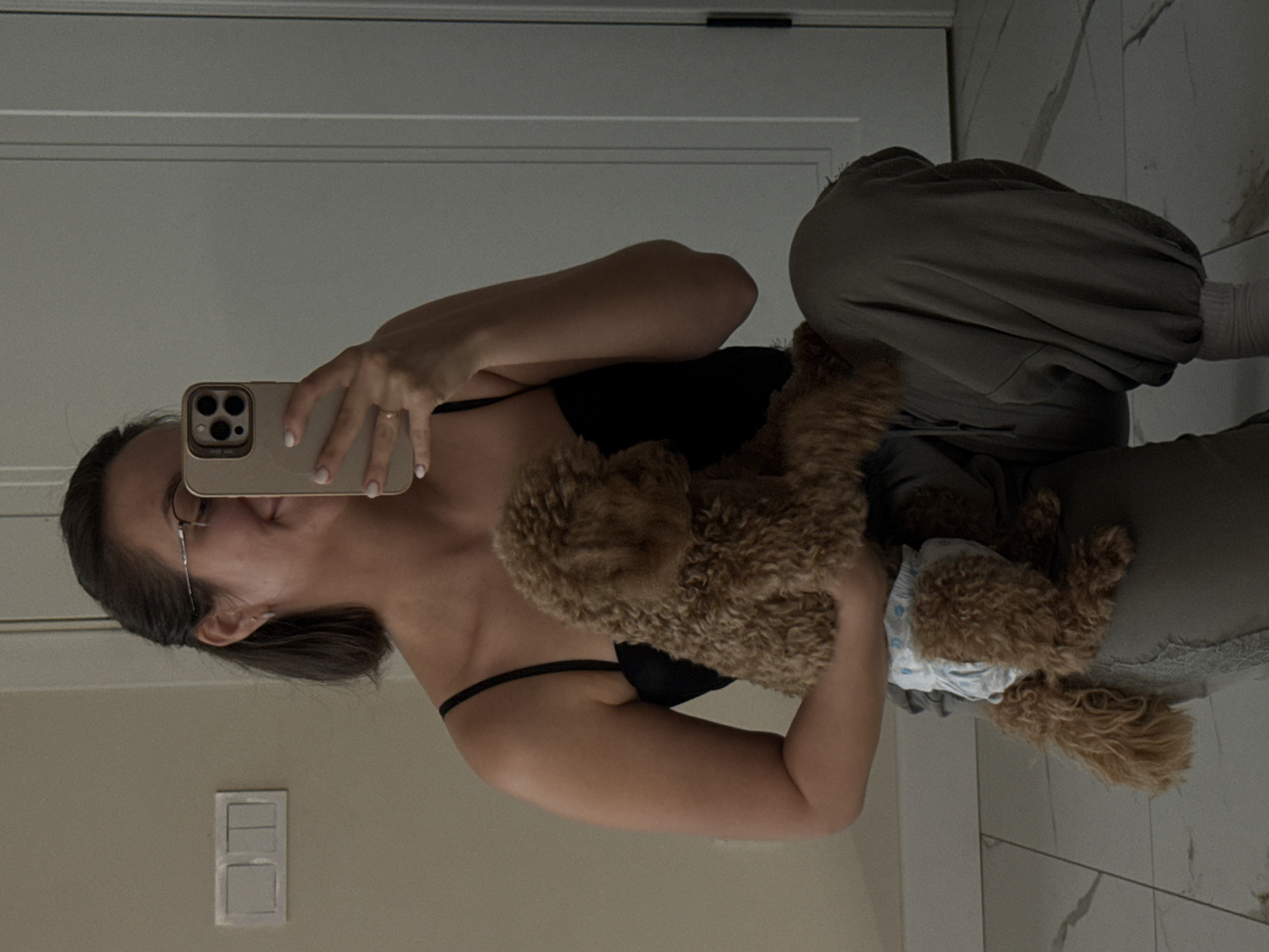 A young adult is kneeling on a tiled floor, holding a small, fluffy brown dog wearing a blue and white patterned diaper. The person is wearing a black tank top and loose-fitting olive green pants, with one foot visible wearing a light pink sock. They are holding a gold-colored smartphone in their right hand, angled as if taking a selfie in a mirror. The person is wearing glasses and has dark hair pulled back. To the left of the person is a white light switch with two rectangular buttons. The background shows a light-colored wall and a partially visible white door on the right. The lighting is soft and appears to be coming from overhead or a nearby window. This scene is not identifiable as being located in Pervomaysky, Russia.