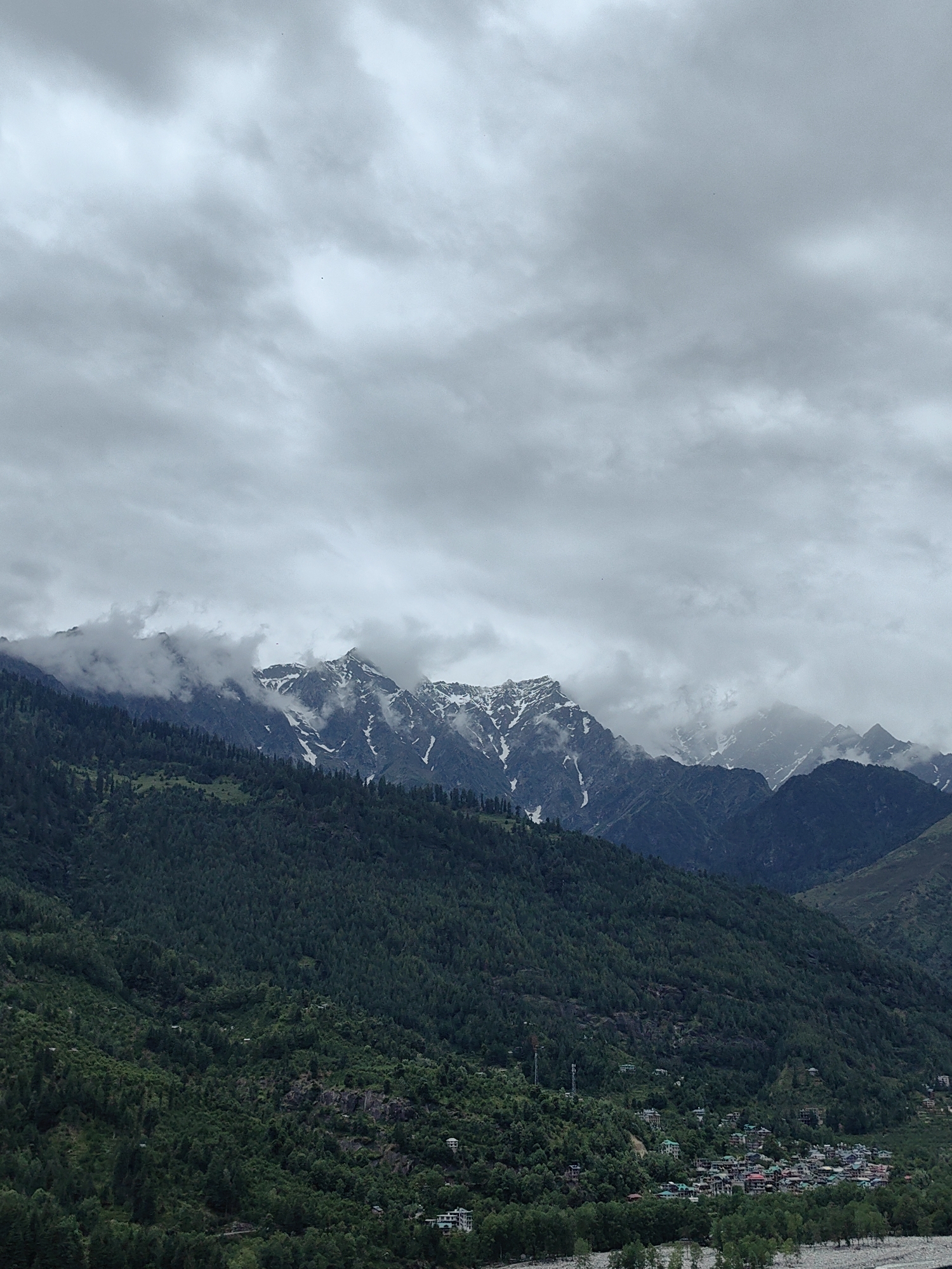 The image depicts a landscape scene in Vashisht, India. Above, there is a cloudy sky with gray hues. Below, in the distance, there are mountain ranges with some visible snow patches on their peaks. The midground features a heavily forested slope, lush with green trees. Further down, a small village with buildings clustered together is visible, located along a riverbank. The overall impression is of a mountainous, verdant landscape with a village nestled within it under overcast conditions.