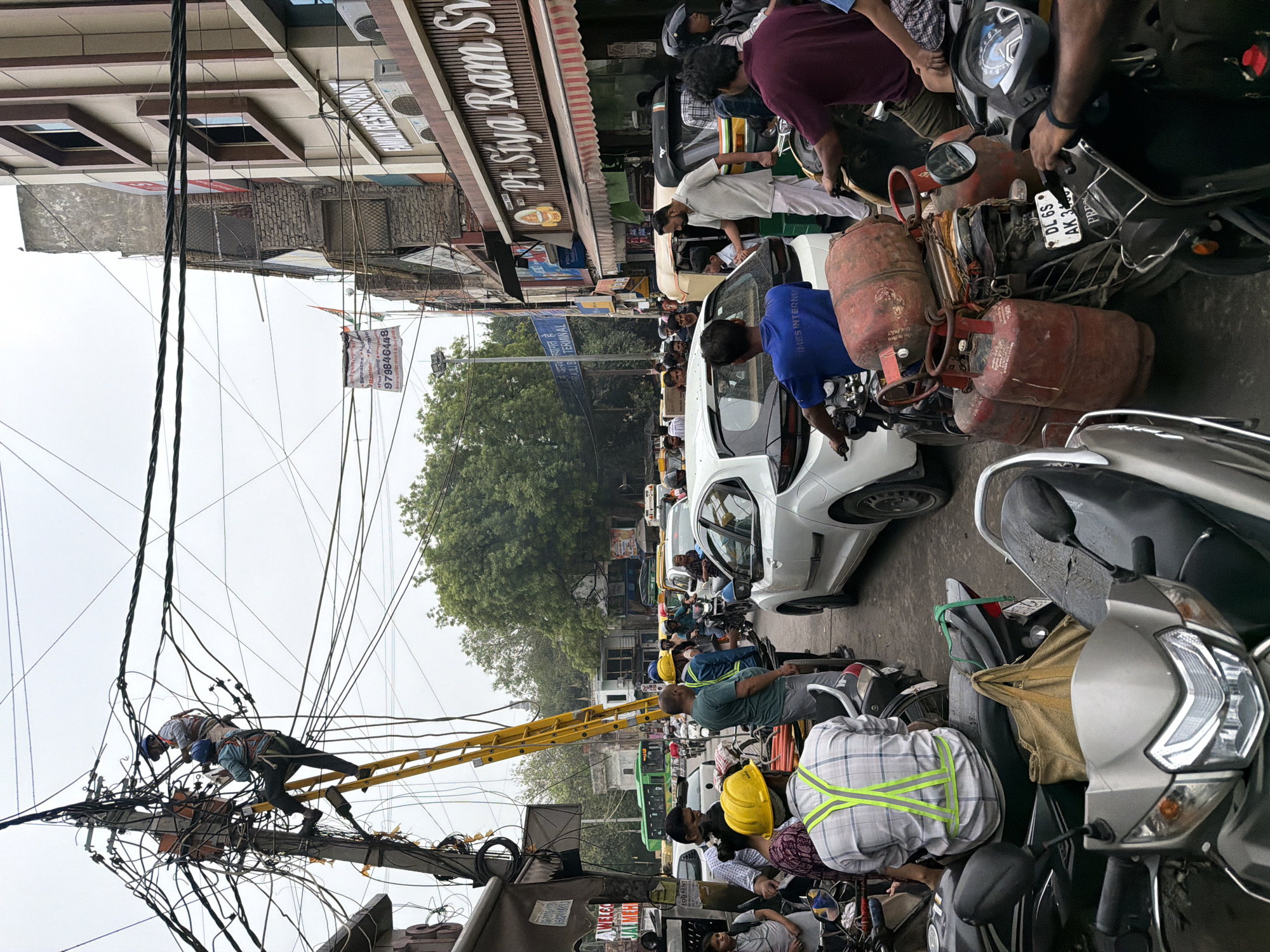 In a crowded street in Delhi, India, two electricians are working on a pole with a multitude of tangled electrical wires and a yellow ladder. One electrician is higher up on the pole, near a transformer, and the other is on the ladder, further down. Below them, a white car is stopped in traffic, with several people on motorcycles and scooters also present. Some of the people are wearing safety vests and hard hats. The street is lined with buildings, one of which has a sign for "Pt. Siya Ram Sweets." Other businesses and structures are visible, along with trees and a green bus in the background. The scene depicts a busy urban environment with ongoing infrastructure work.