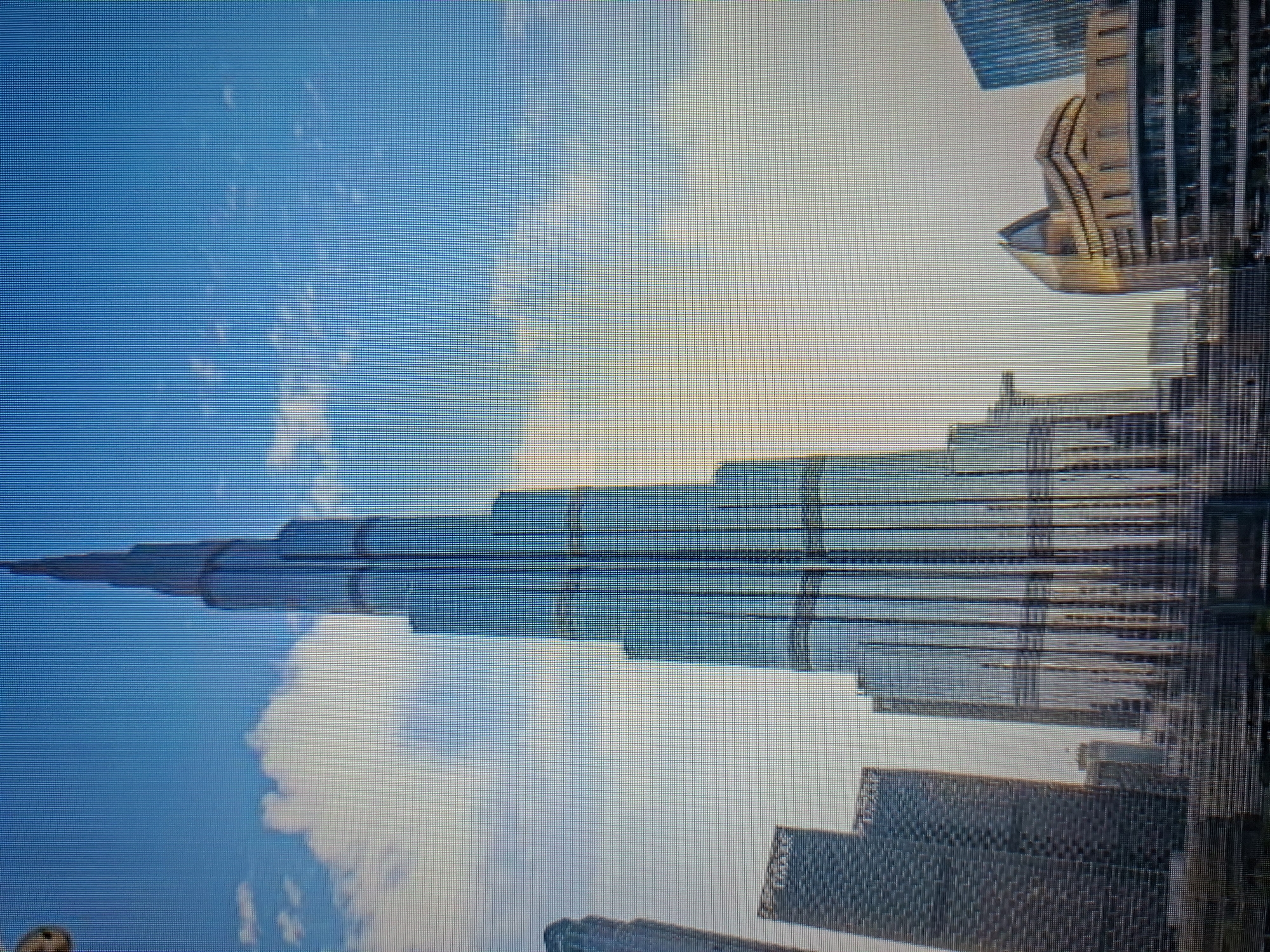 The image depicts the Burj Khalifa skyscraper in Dubai, United Arab Emirates. The tower stands tall against a backdrop of blue sky with some clouds. Other buildings are visible around the base of the Burj Khalifa.
