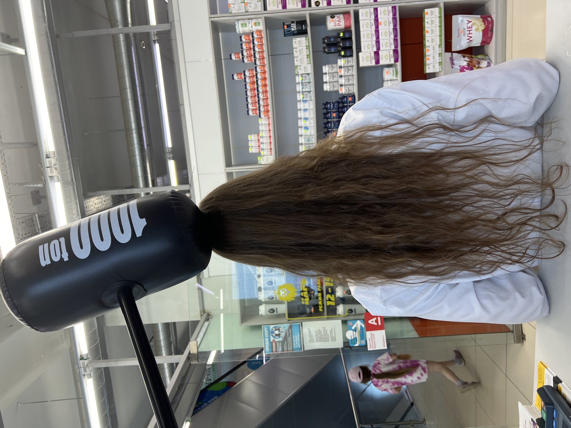 A woman with long brown hair, viewed from behind, stands facing away from the camera, wearing a white lab coat. A large, black, inflatable hammer with "1000 ton" written in white on its side rests atop her head. The hammer is attached to a long black handle. The scene appears to be an interior of a store, possibly a pharmacy or health supplement shop, with shelves stocked with bottles and packages of various products in the background. To the left, a child wearing a pink tie-dye shirt and a pink baseball cap walks away from the camera, near an escalator. The overall lighting suggests an indoor commercial setting. The location is identified as Chaikovsky, Russia.