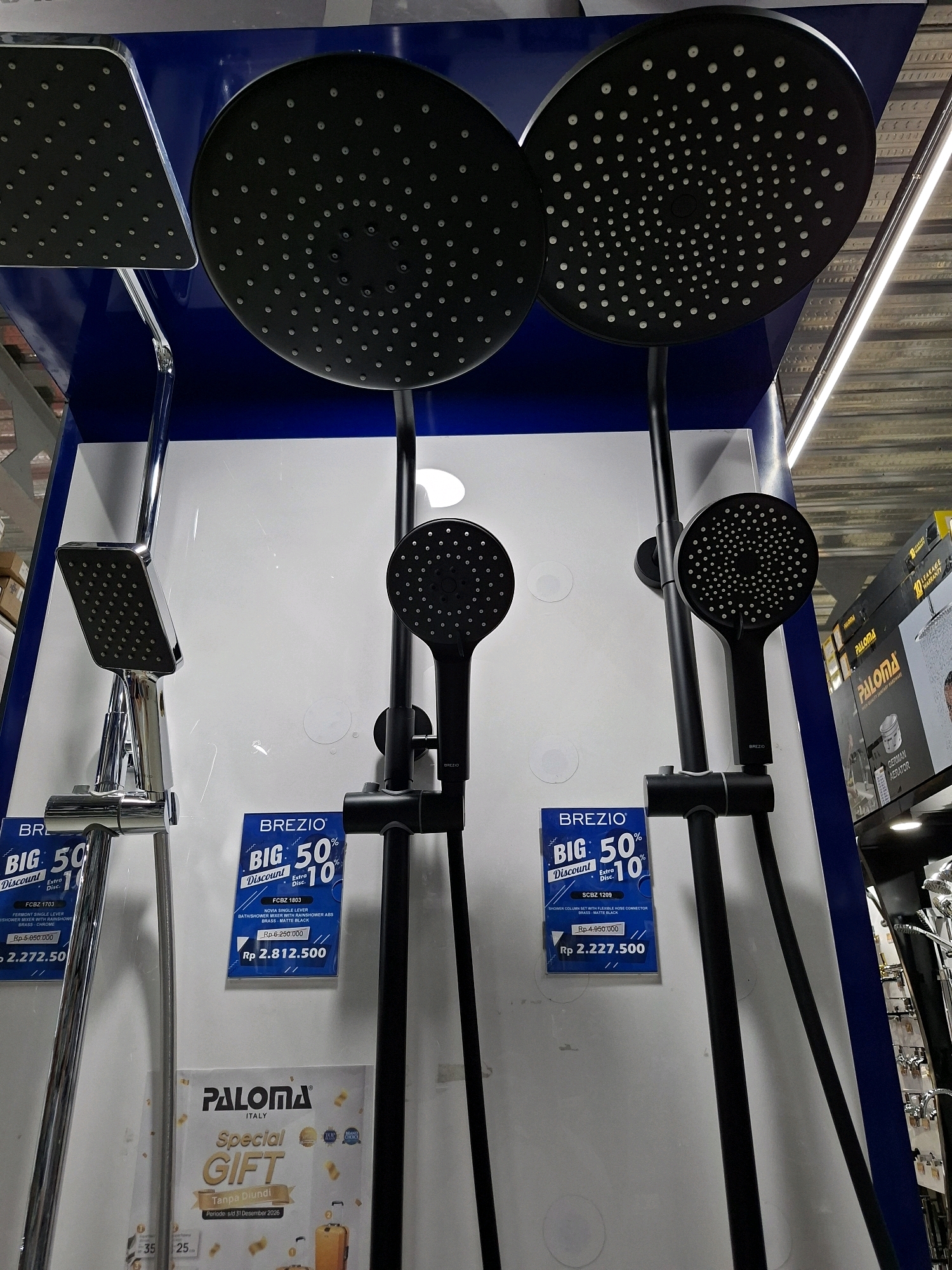 The image displays a retail environment, likely a bathroom fixtures store in South Tangerang, Indonesia. The focal point is a display of modern shower heads and mixer systems. On the left, a chrome-finished square shower head is attached to a mixer. To its right, a black handheld shower head is mounted on a black shower column. Further to the right, another black handheld shower head is visible on a similar black shower column. Two prominent blue and white price tags are attached to the display. One tag reads "BREZIO BIG 50% Discount Extra Disc. 10% Rp 2.812.500". Another tag, partially obscured, also mentions "BREZIO BIG 50%". Below the shower displays, a promotional flyer with "PALOMA ITALY Special GIFT Tanpa Di Undi" is visible. The background shows a blue wall and a white display surface. In the upper right portion of the image, a section of shelving with various bathroom fixtures and packaging is visible, including items with "PALOMA" branding. The overall impression is a well-lit retail setting showcasing plumbing fixtures.