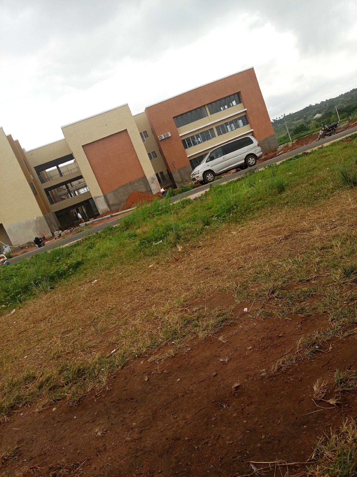 The image shows a modern, multi-story building under construction in Iseyin, Nigeria. The building features a beige and orange facade with multiple windows. A silver minivan is parked in front of the building. Construction materials, including mounds of dirt and scaffolding, are visible. The surrounding area consists of a grassy field and a dirt road. The sky is overcast with clouds. Several individuals are present, seemingly engaged in construction activities.