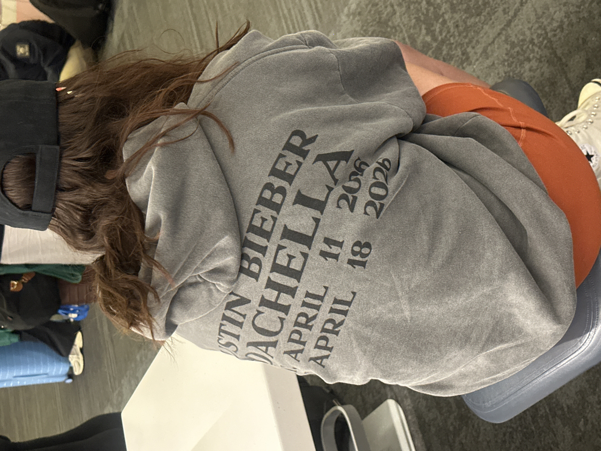 A person wearing a gray hoodie, orange shorts, and a black baseball cap is seated in an interior space. The hoodie has "Justin Bieber Coachella" printed on the back, along with "April 11 2026" and "April 18 2026". Luggage is partially visible in the lower left corner. The person is sitting on a blue chair. The location is in the city of Los Angeles, United States of America.