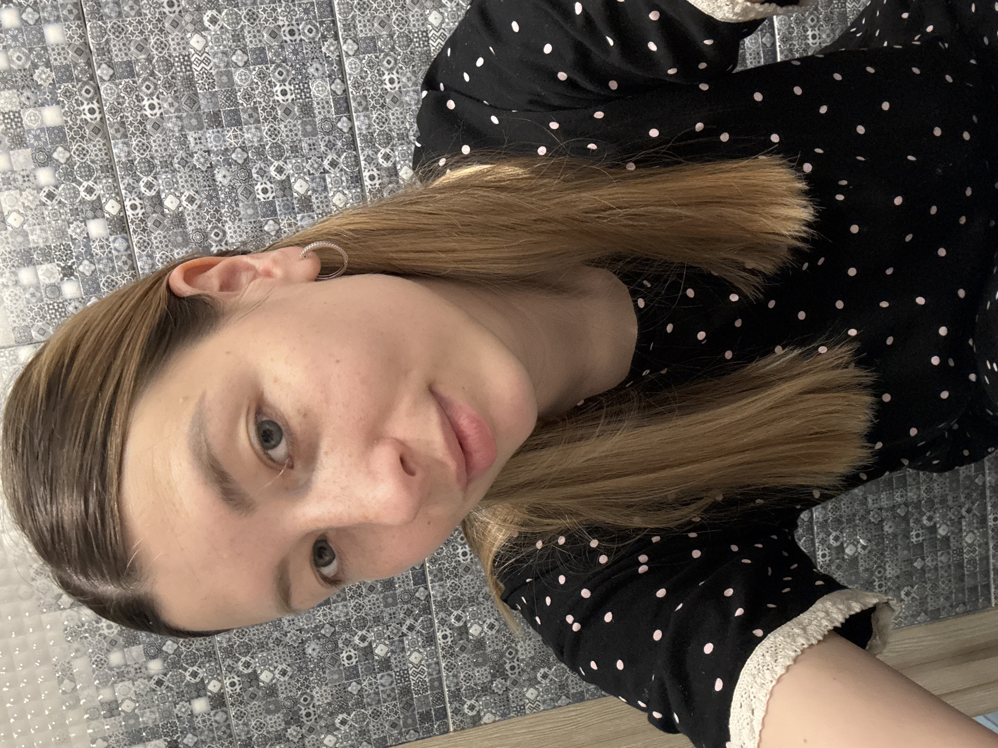 The image depicts a young woman with fair skin, light brown hair, and light-colored eyes. She is wearing a black garment with pink polka dots and white lace trim. An earring is visible in her left ear. The background consists of patterned tiles. There is no indication of this photo being in Moscow, Russia. The only person visible is the young woman.