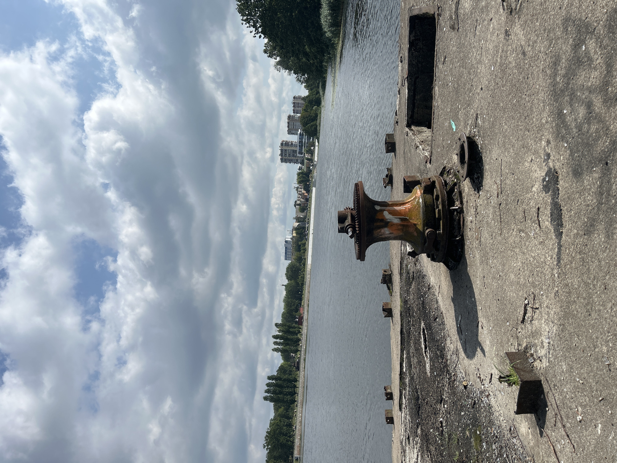 The image depicts a waterfront scene in Brest, Belarus, likely a disused industrial or port area. In the foreground, a weathered, rusty winch mechanism is centrally positioned on a concrete pier. The pier is rough and weathered, with patches of debris and several square metal fixtures embedded in the concrete. The water of a large river or canal stretches out behind the pier, appearing calm with ripples on its surface. Across the water, a tree-lined embankment is visible, with several tall, slender trees standing out against a backdrop of distant buildings, including apartment complexes and modern architectural structures. The sky above is filled with a mix of white and gray clouds, suggesting overcast or partially cloudy weather. The overall impression is one of industrial decay and the reclaiming of space by nature, with a city skyline in the distance.