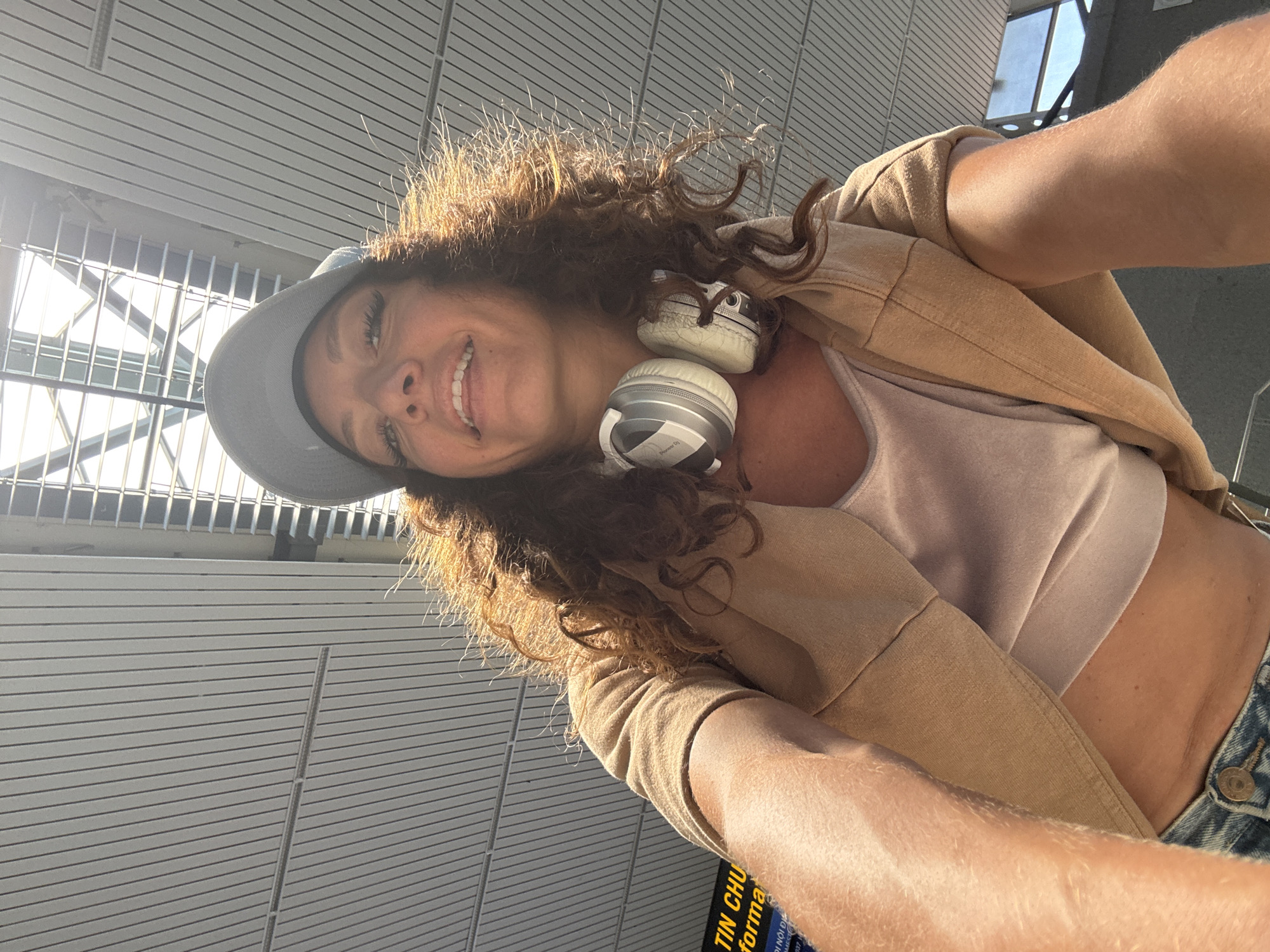 A woman takes a selfie in an airport. She wears a grey baseball cap, a tan jacket over a light pink top, and blue jeans. Silver headphones rest around her neck. The background includes a white, horizontally-lined wall and a window with metal bars. A sign with Vietnamese text is partially visible in the lower left corner. The photograph appears to be taken in the airport of Đà Nẵng, Vietnam.