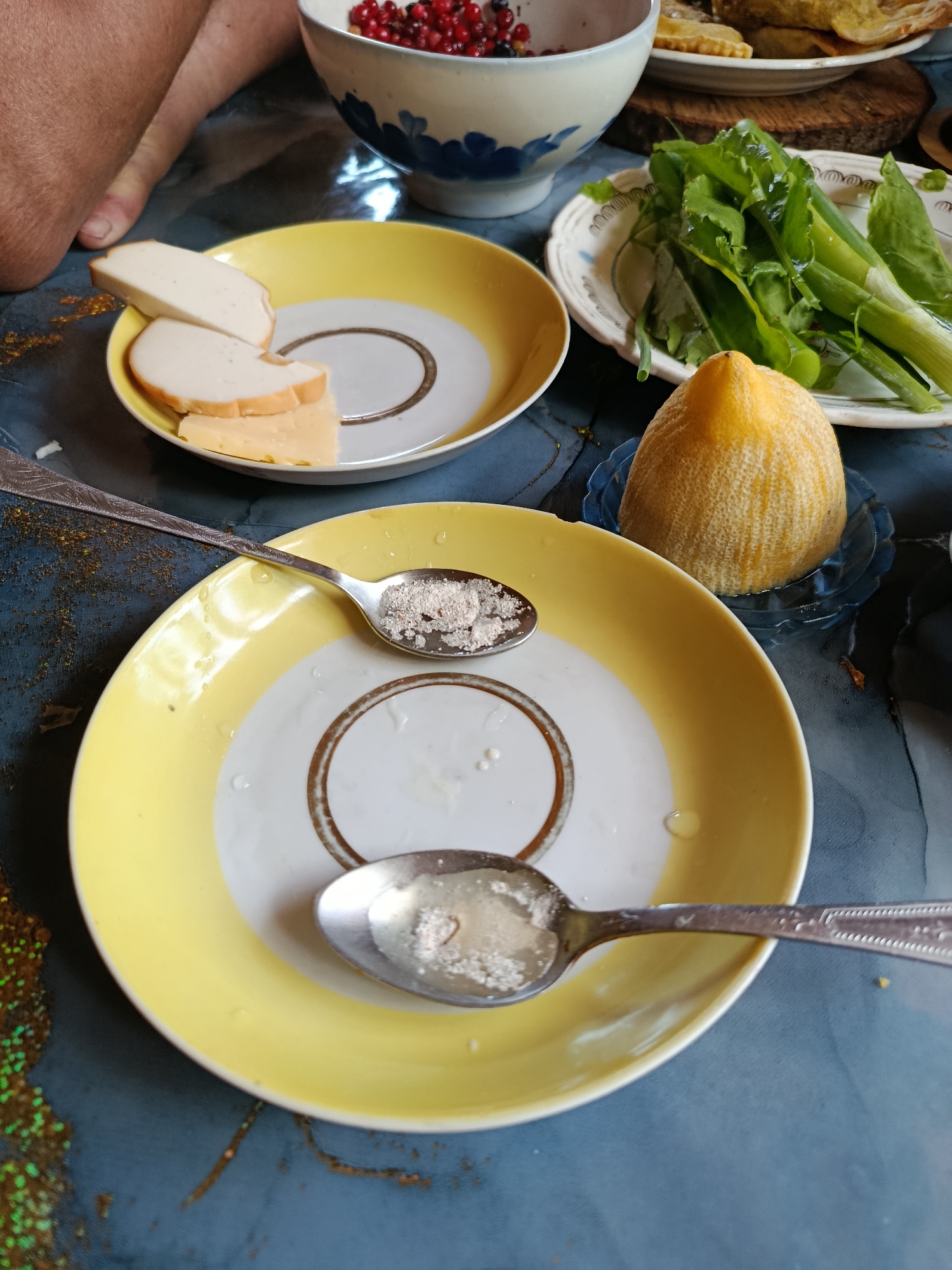 A close-up, overhead view depicts a domestic tabletop scene, likely during or after a meal, in the city of Unknown, Unknown.
In the upper left quadrant, a portion of a human arm and elbow, with light-toned skin, rests on a dark blue table surface. Adjacent to the arm, a small scattering of iridescent golden-brown granular material is visible on the tabletop.
Towards the upper right, a white ceramic bowl with blue decorative motifs is partially visible, containing red berries (resembling cranberries or redcurrants) and some darker fruit or seeds. Behind this bowl, a stack of golden-brown baked goods, possibly pastries or flatbreads, rests on a white plate, which in turn sits on a circular wooden trivet or board.
In the mid-left foreground, a round white plate with a yellow rim and a central brown concentric circle design holds several slices of food. Two crescent-shaped slices of a pale, uniform substance (possibly processed meat or cheese) are layered, with a third, yellowish, more irregularly shaped slice of cheese positioned beneath them.
In the mid-right, a white, irregularly shaped ceramic plate with a faint patterned rim holds a cluster of fresh green leafy vegetables and several stalks of green onions. These greens appear to have a sheen of moisture or oil. In front of this plate, a whole, unpeeled lemon with a textured, slightly ribbed rind is placed in a small, fluted, clear blue glass dish.
In the lower-center, another round white plate identical to the one in the mid-left foreground is present. This plate contains several droplets of clear liquid. Two silver-colored metal spoons rest on this plate. The upper spoon contains a small mound of light brown, granular powder. The lower spoon, which has an embossed pattern on its handle, also contains a smaller quantity of similar light brown powder.
The table surface itself is a dark blue with lighter blue or white marbling, appearing somewhat glossy. Scattered across the table, particularly in the lower left, are minute specks of green and golden glitter-like material.