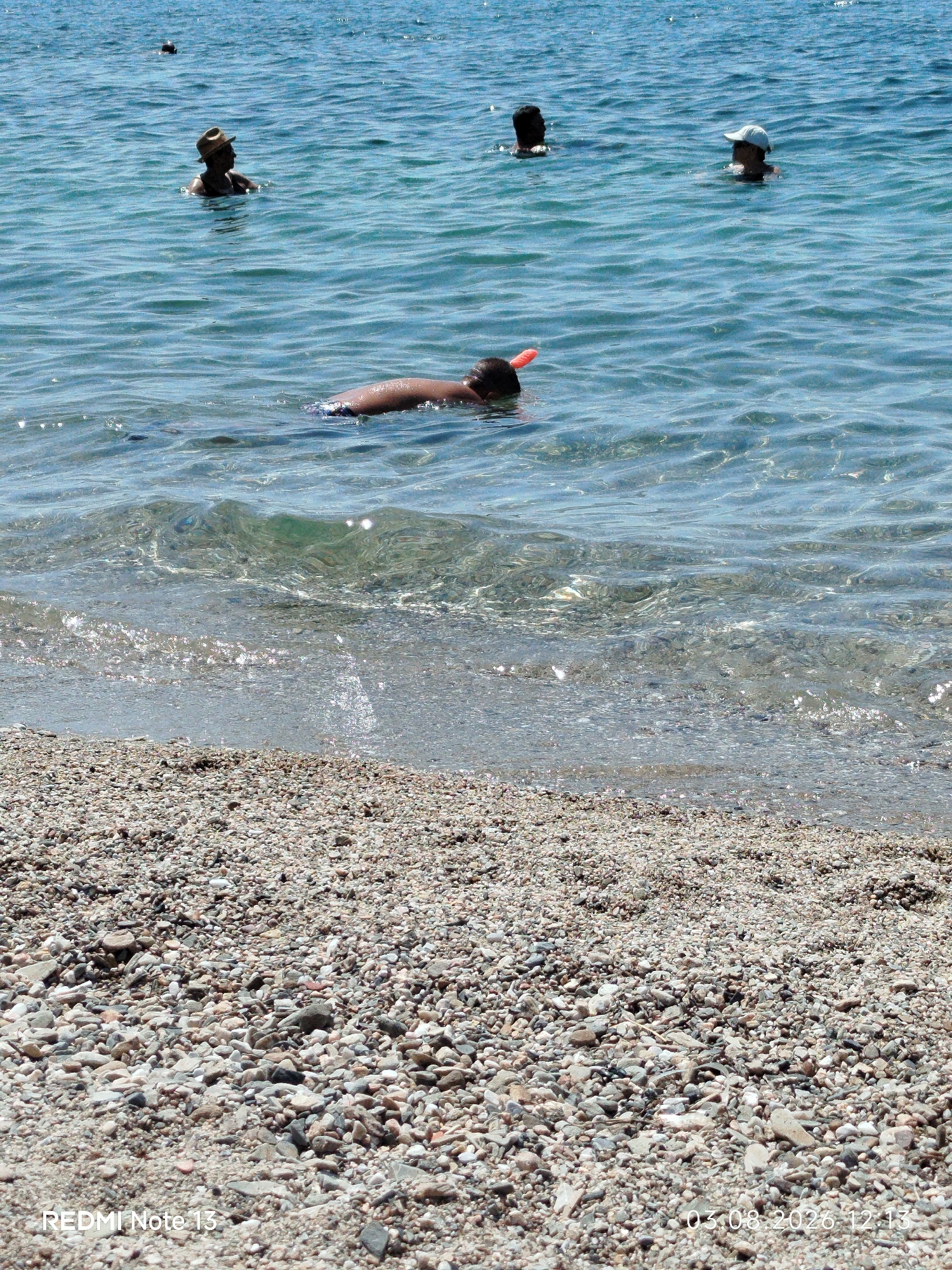 The image shows a group of people in the clear blue waters of a beach in Sithonia, Greece. In the foreground, the pebbled shoreline meets the shallow, sparkling water. A person is partially submerged, wearing a snorkel mask with an orange mouthpiece, appearing to be swimming or exploring beneath the surface. Further out in the water, other individuals are visible, some wearing hats. The sun is shining, creating reflections on the water's surface. The scene depicts a typical summer day at a Greek beach.