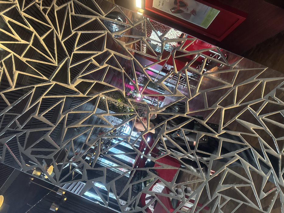 A fragmented reflection of an individual taking a self-portrait with a smartphone is captured in a decorative mirror within an indoor commercial establishment in Tbilisi, Georgia. The mirror's surface is composed of numerous irregular polygonal segments (triangles and quadrilaterals) separated by dark, geometric grid lines, creating a fractured visual effect. The reflected person, visible from the mid-torso up, wears a dark leather jacket over dark apparel and has distinct red lipstick. The smartphone is held at arm's length, angled towards the individual.
The reflected environment behind the person indicates a modern indoor setting, likely a restaurant or cafe. Visible elements include red-upholstered tables or chairs, a prominent red wall or partition, and a framed poster or sign partially visible on the right. The sign displays text beginning with "How did..." and "make a..." along with an image of a person wearing red heart-shaped sunglasses. Overhead light fixtures emitting a warm yellow glow are reflected, as are sections of a dark, slatted ceiling. Areas within the reflection show ambient pink and magenta lighting.