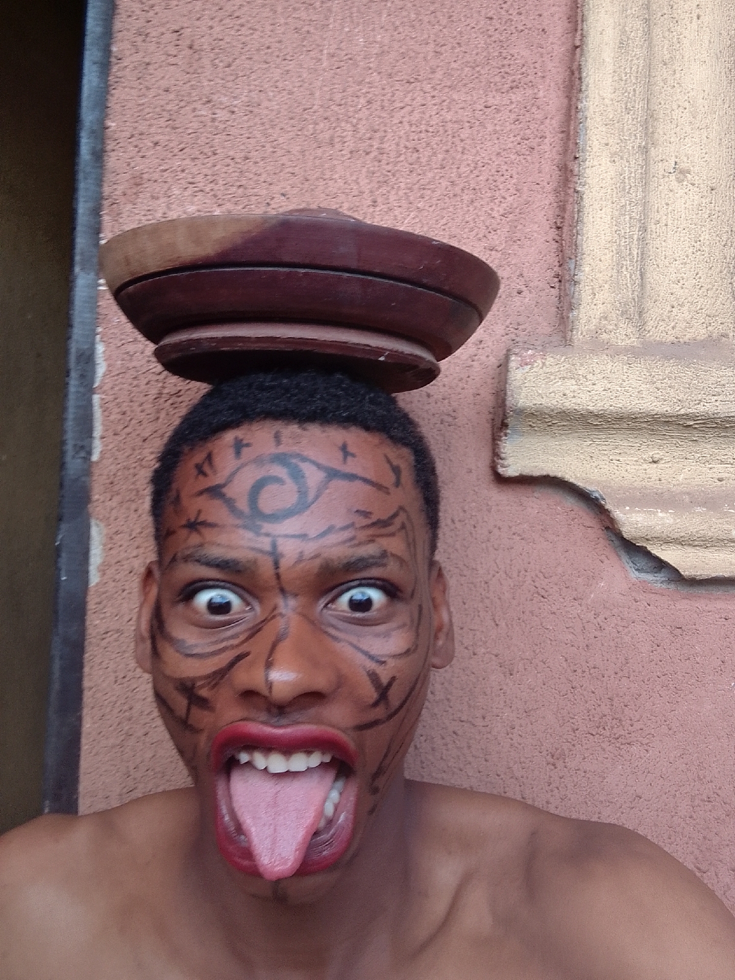 A close-up shot depicts a topless man with dark skin, likely in his late teens or early twenties, wearing black face paint resembling tribal or ritualistic markings. An eye is drawn on his forehead, surrounded by other symbols. He wears bright red lipstick and his mouth is wide open, revealing his teeth and tongue. Atop his head, he balances a wooden bowl or container. The background consists of a textured, pinkish-orange wall with a partial architectural feature resembling a corner molding to the right. The image seems to be taken in an urban location. The specific location and city are not discernible from the image.